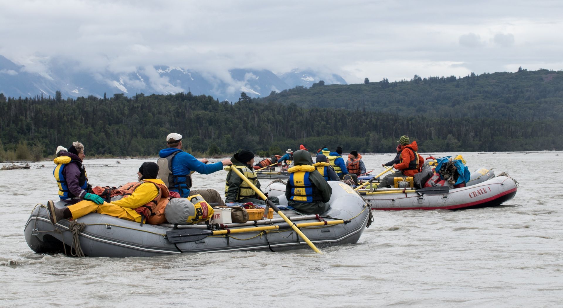 People rafting on a river, with mountains and trees in the background under a cloudy sky.