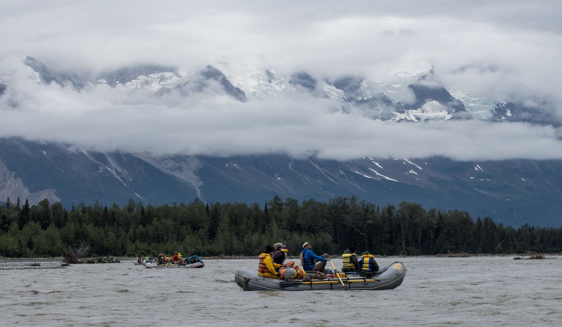 Rafts on a river with people, cloudy mountain backdrop, and green trees lining the shore.