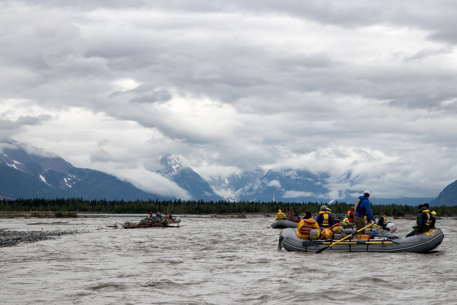 Rafts with people on river, mountains in background under cloudy sky.