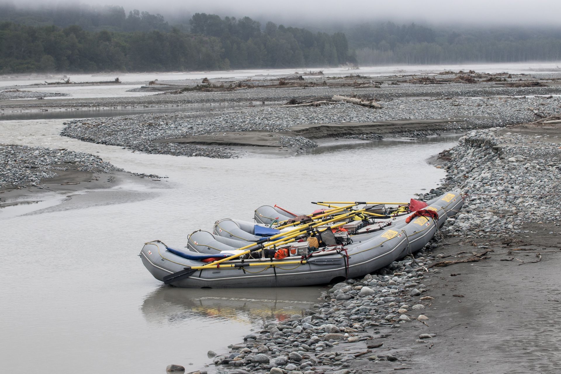 Rafts pulled ashore on a gravel riverbank. Gray sky, silty water, logs in the background.