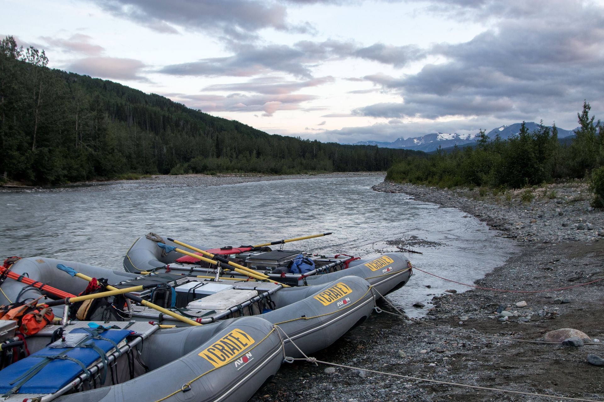 Rafts pulled ashore on a rocky riverbank, overlooking a flowing river and mountain range under a cloudy sky.
