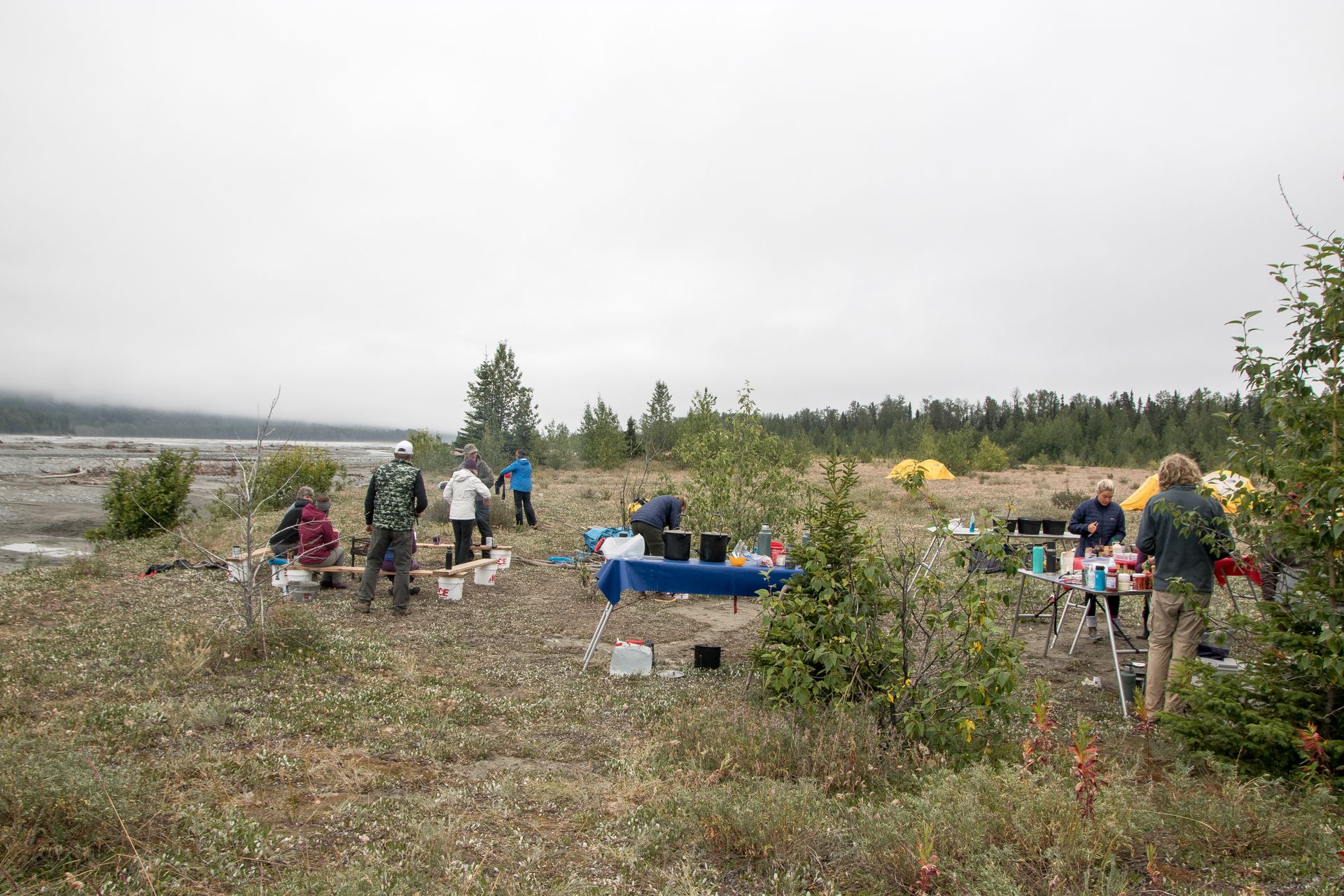 People set up camp by a river, near a forest. Overcast day.