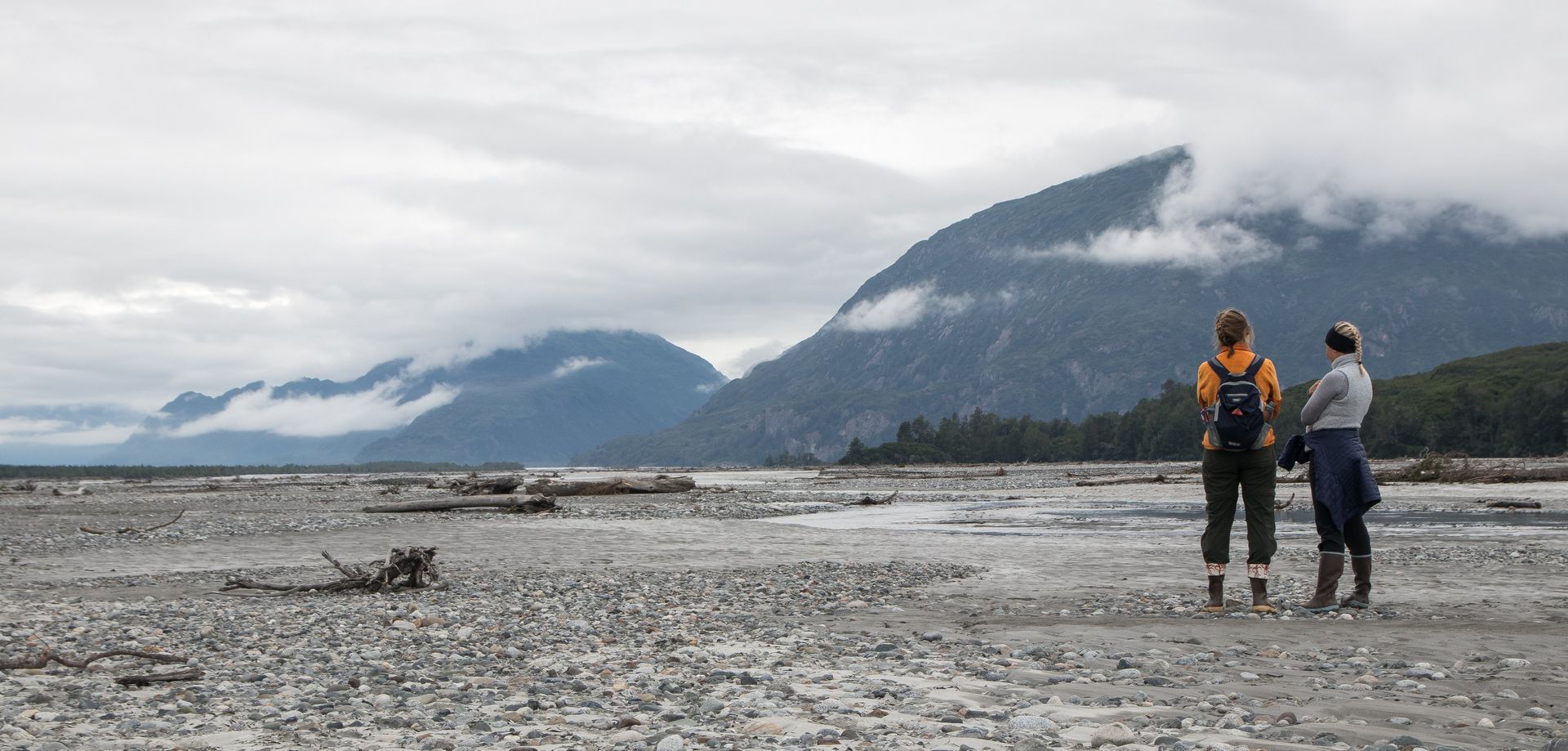 Two people stand on a rocky beach, facing misty mountains under an overcast sky.