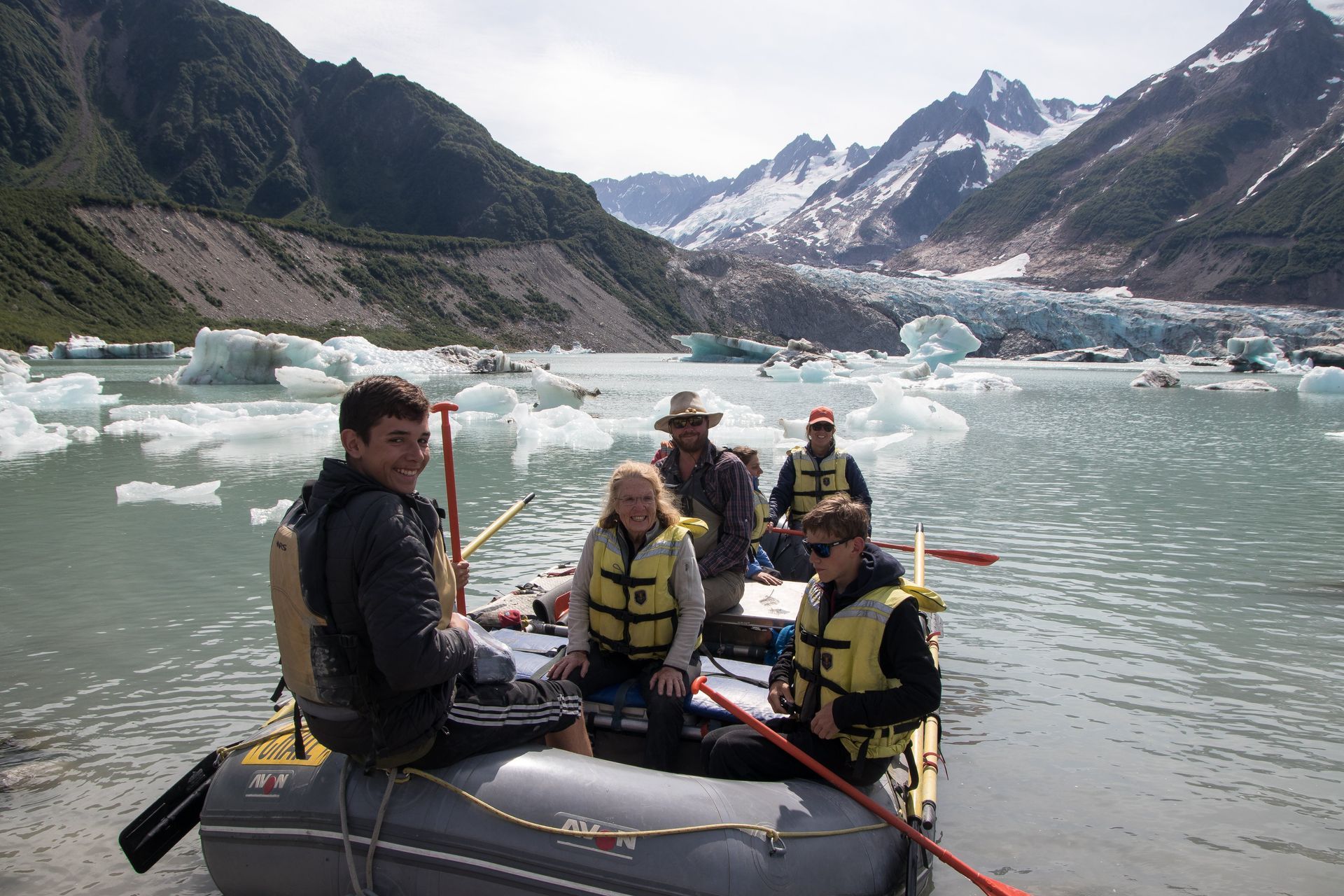People on a raft in a glacial lake, icebergs and mountains in the background.