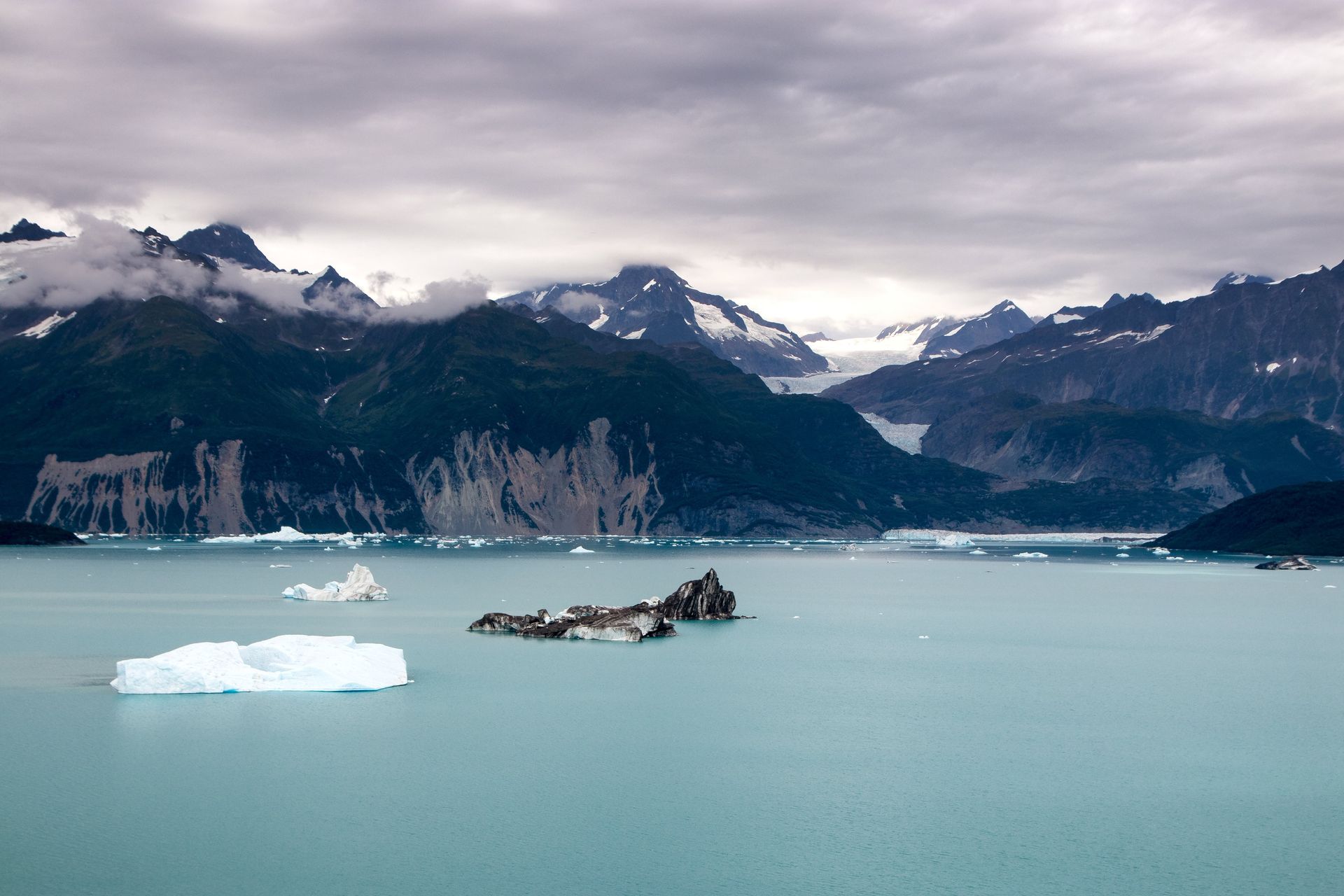 Mountains with snow-capped peaks overlook an ocean with icebergs; overcast sky.