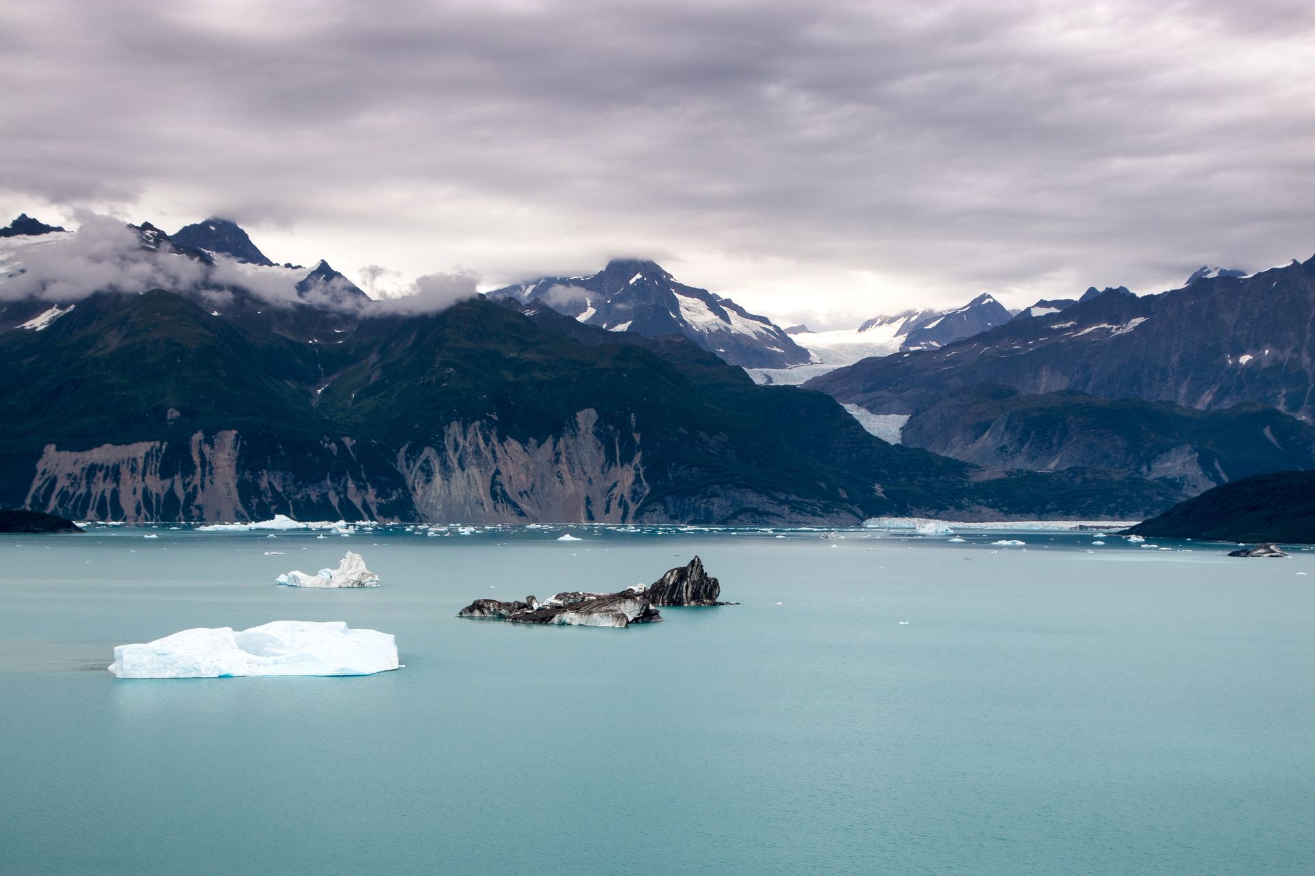 Glacial lake with icebergs, mountains, and overcast sky.