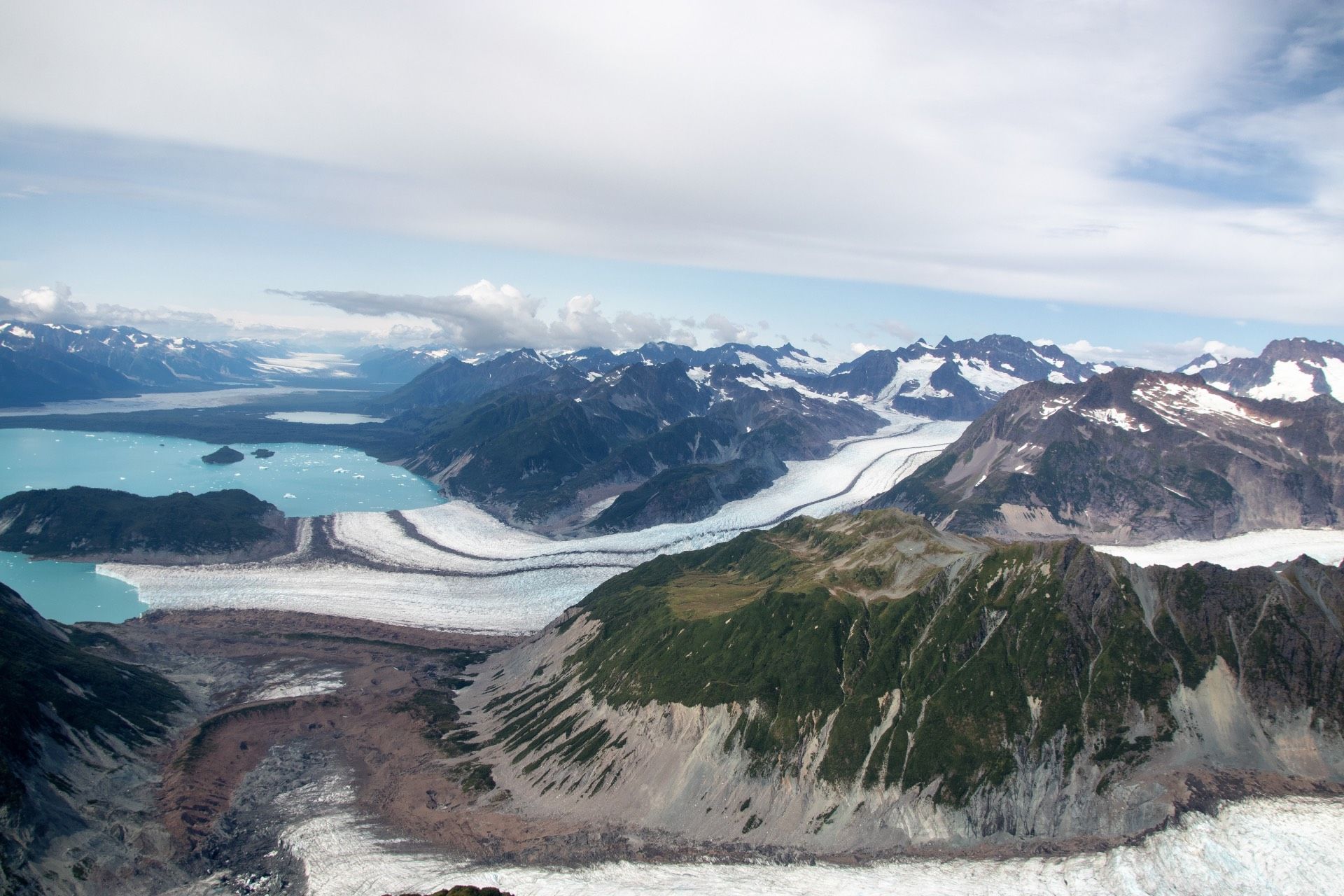 Aerial view of a glacial landscape with turquoise lake, ice, mountains, and green vegetation.