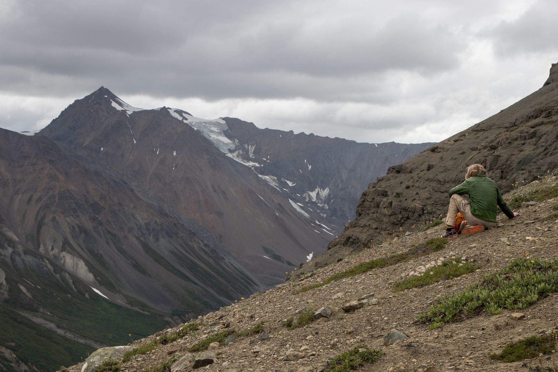 Person seated on a rocky mountainside, looking at a distant, snow-capped peak under a cloudy sky.