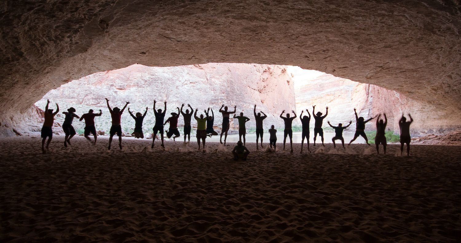 Silhouetted people with arms raised, jumping and cheering in a cave opening.