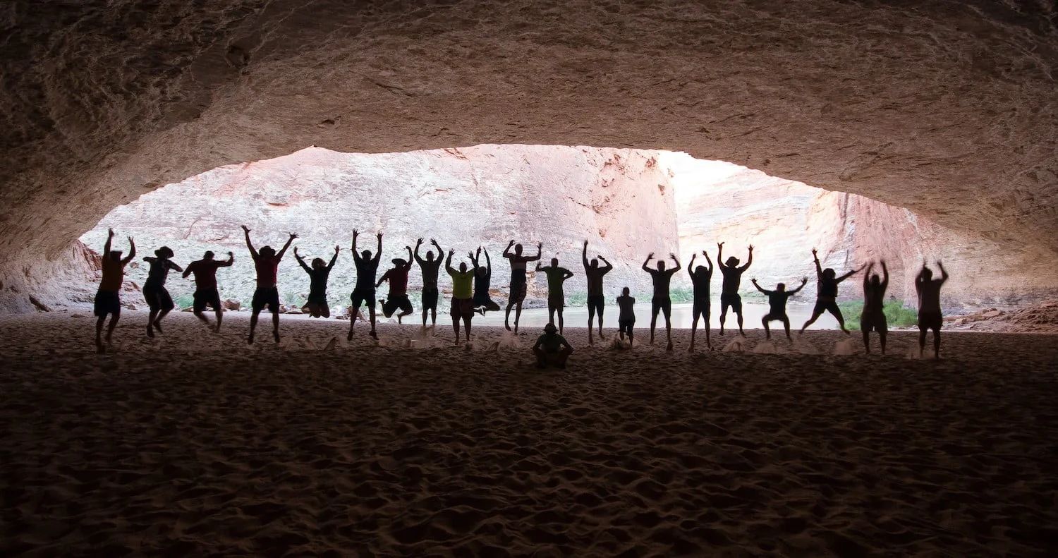 Silhouetted people with arms raised in a cave opening; sand floor, light from above.