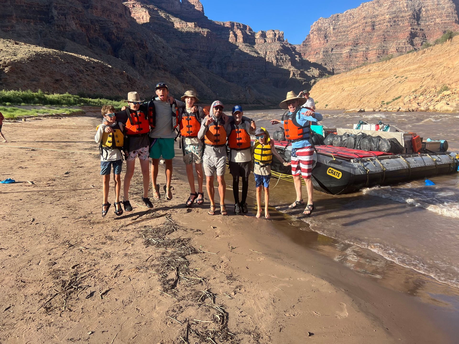 Group of people in life vests pose on a sandy riverbank with a raft and canyon backdrop.