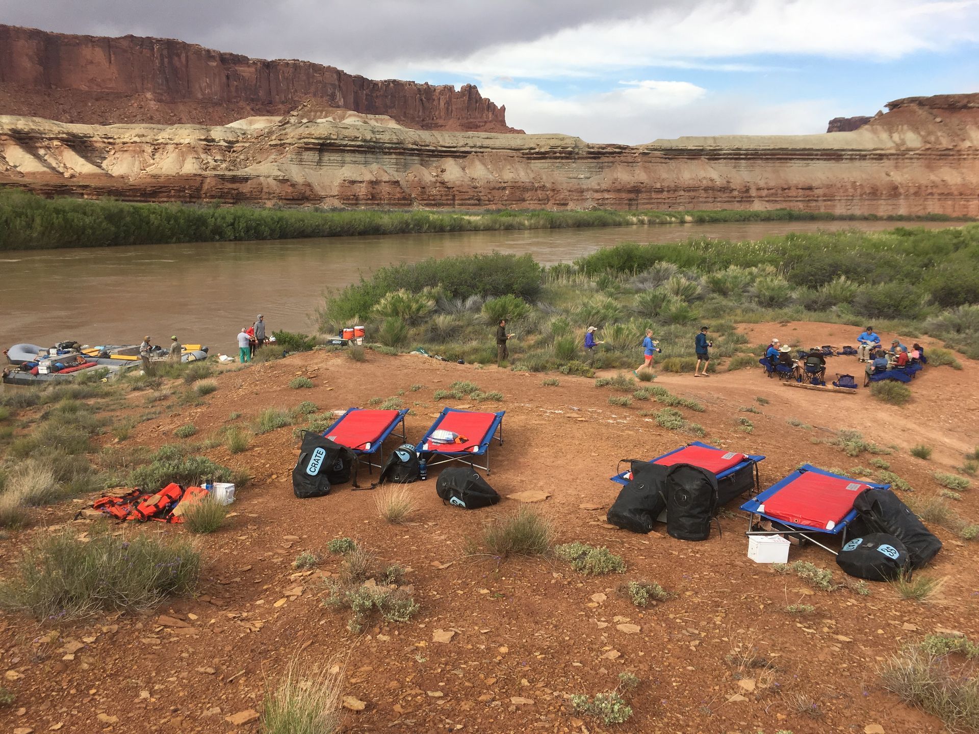 Rafts and gear on a red dirt bank beside a river, people in the background, red rock cliffs.