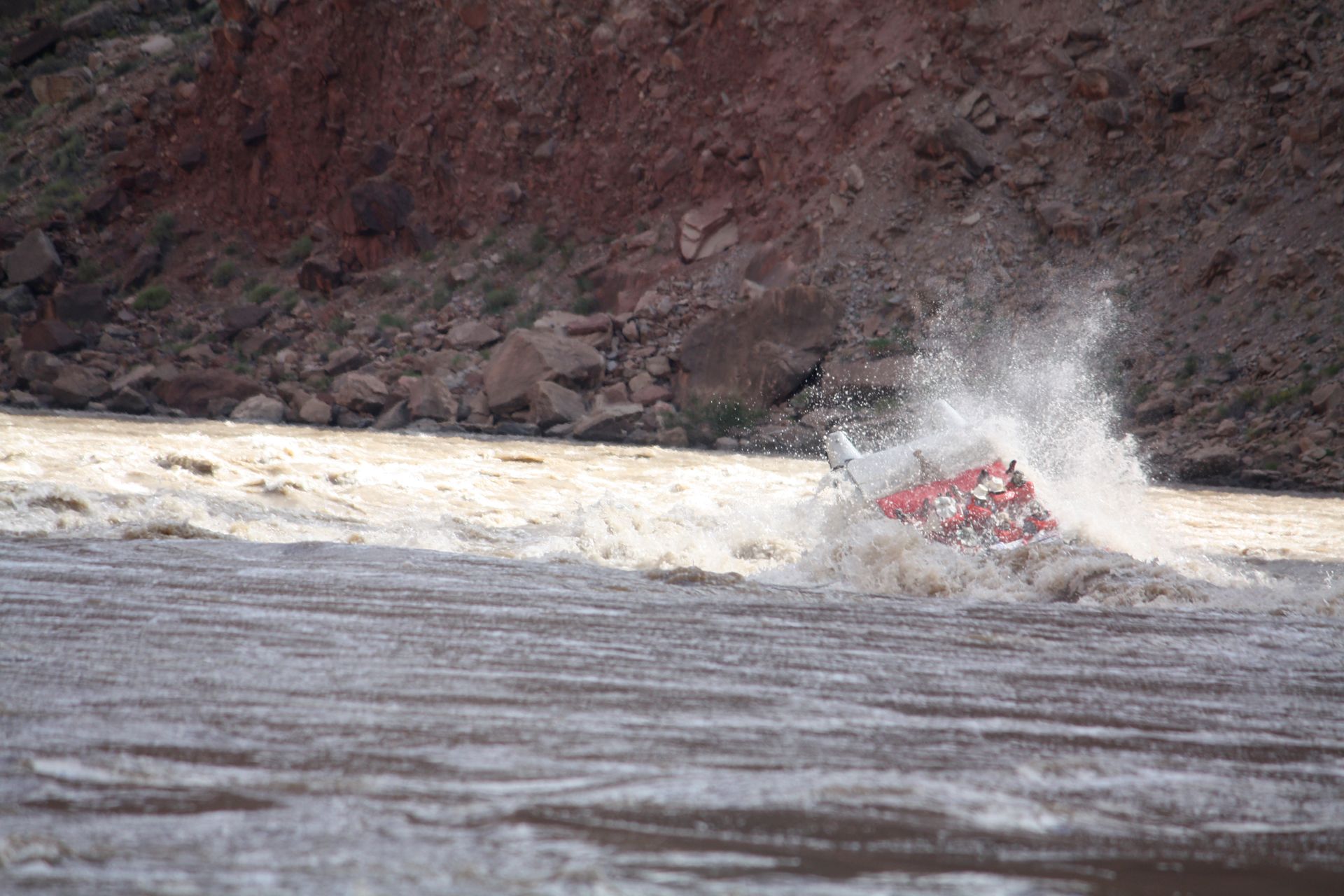 Whitewater raft capsizes in a churning river, creating a large splash. Red canyon walls in background.