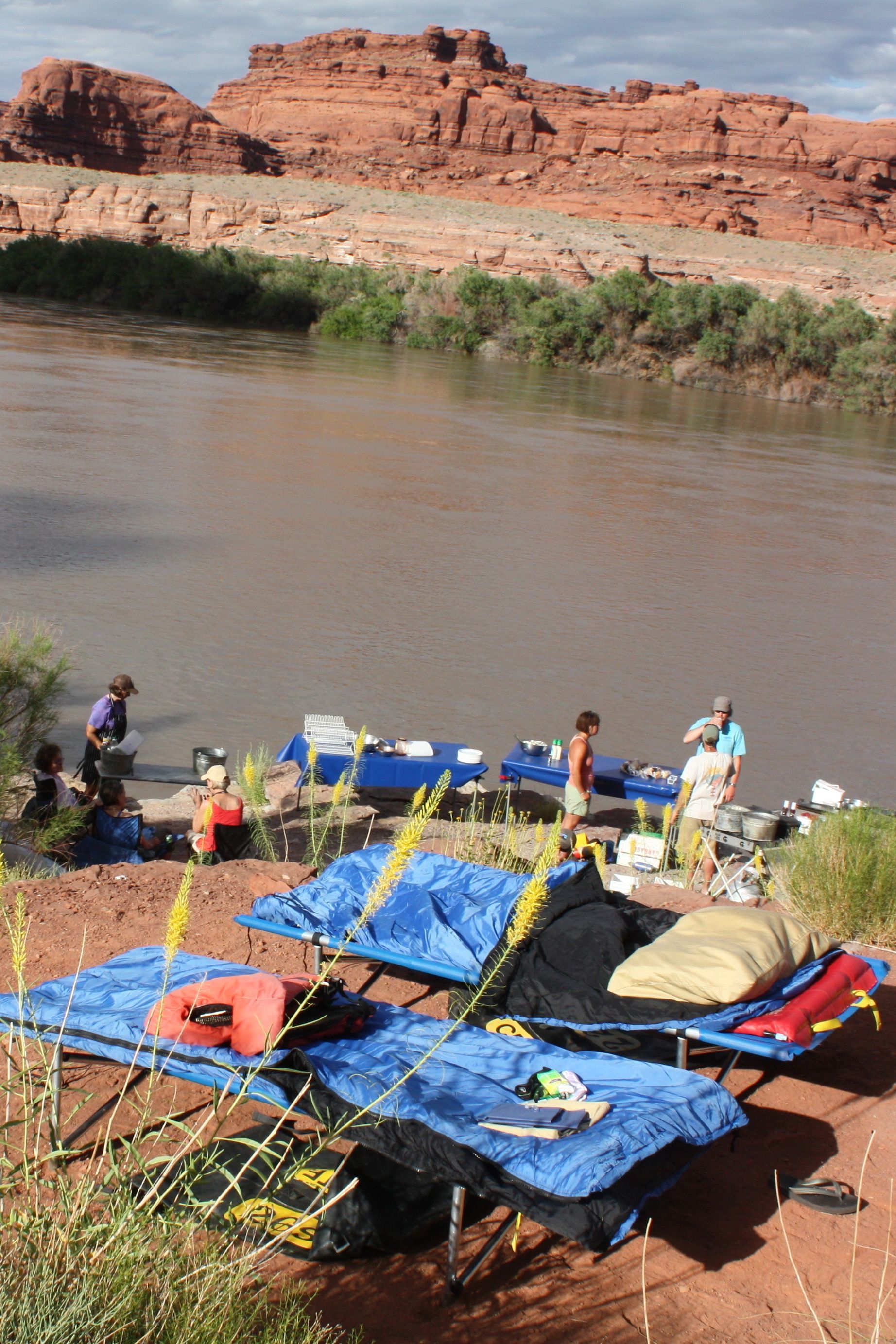 Campers beside a river, setting up cots and supplies with red rock cliffs in the background.