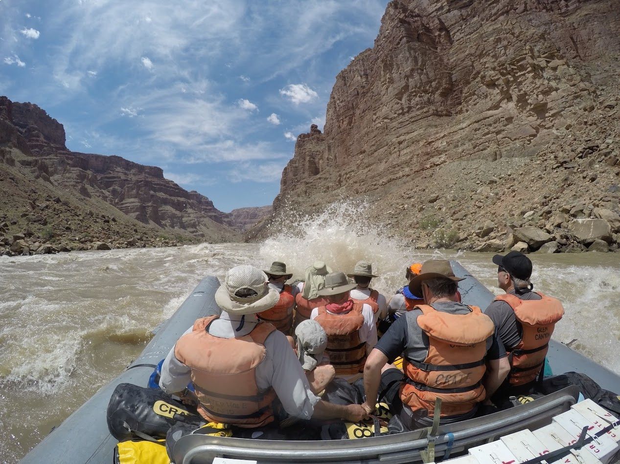 Rafting through a canyon, passengers in life vests get splashed by water. Rocky walls surround the boat.