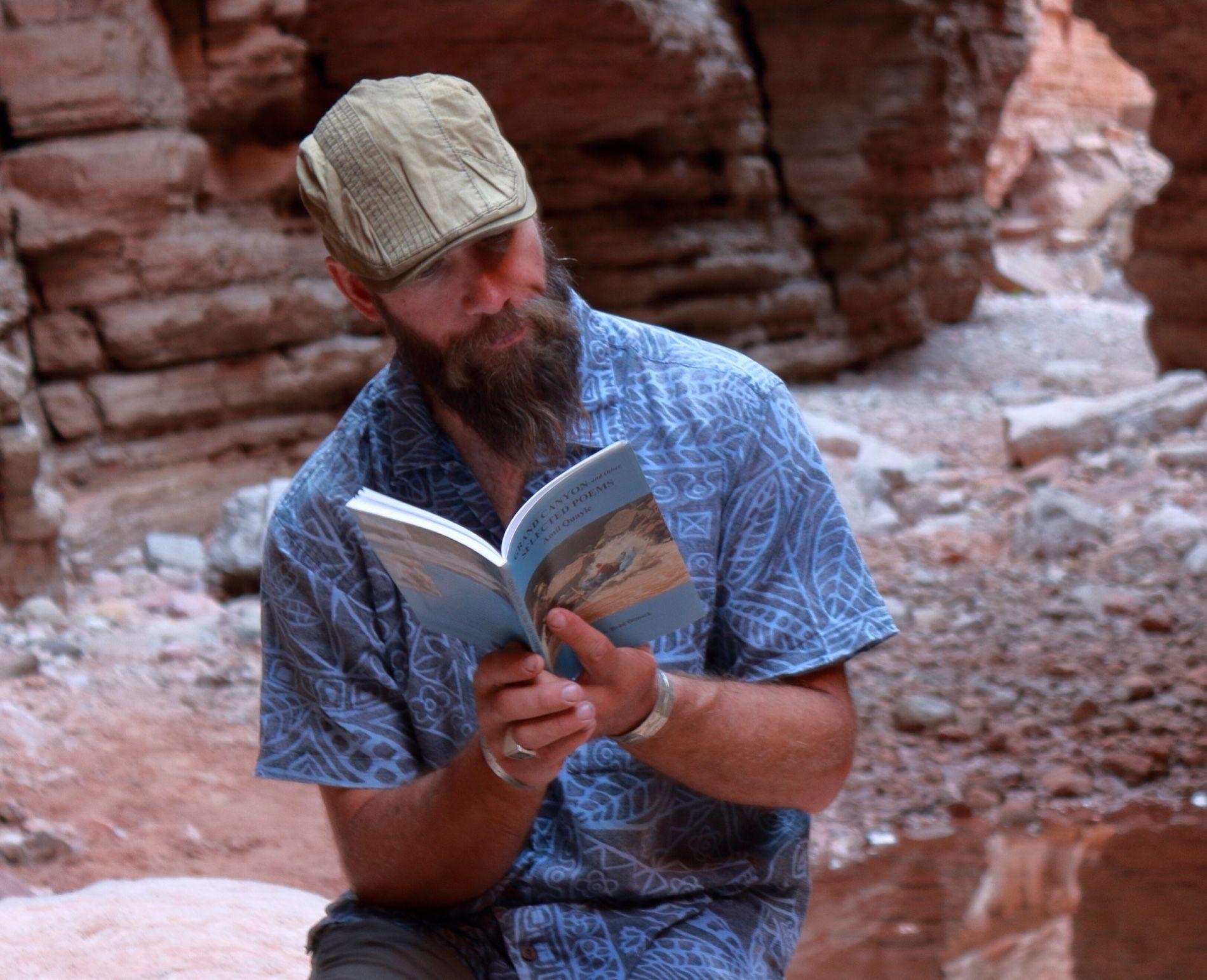 Man with beard reads a book in a red-rock canyon. He wears a cap and blue patterned shirt.