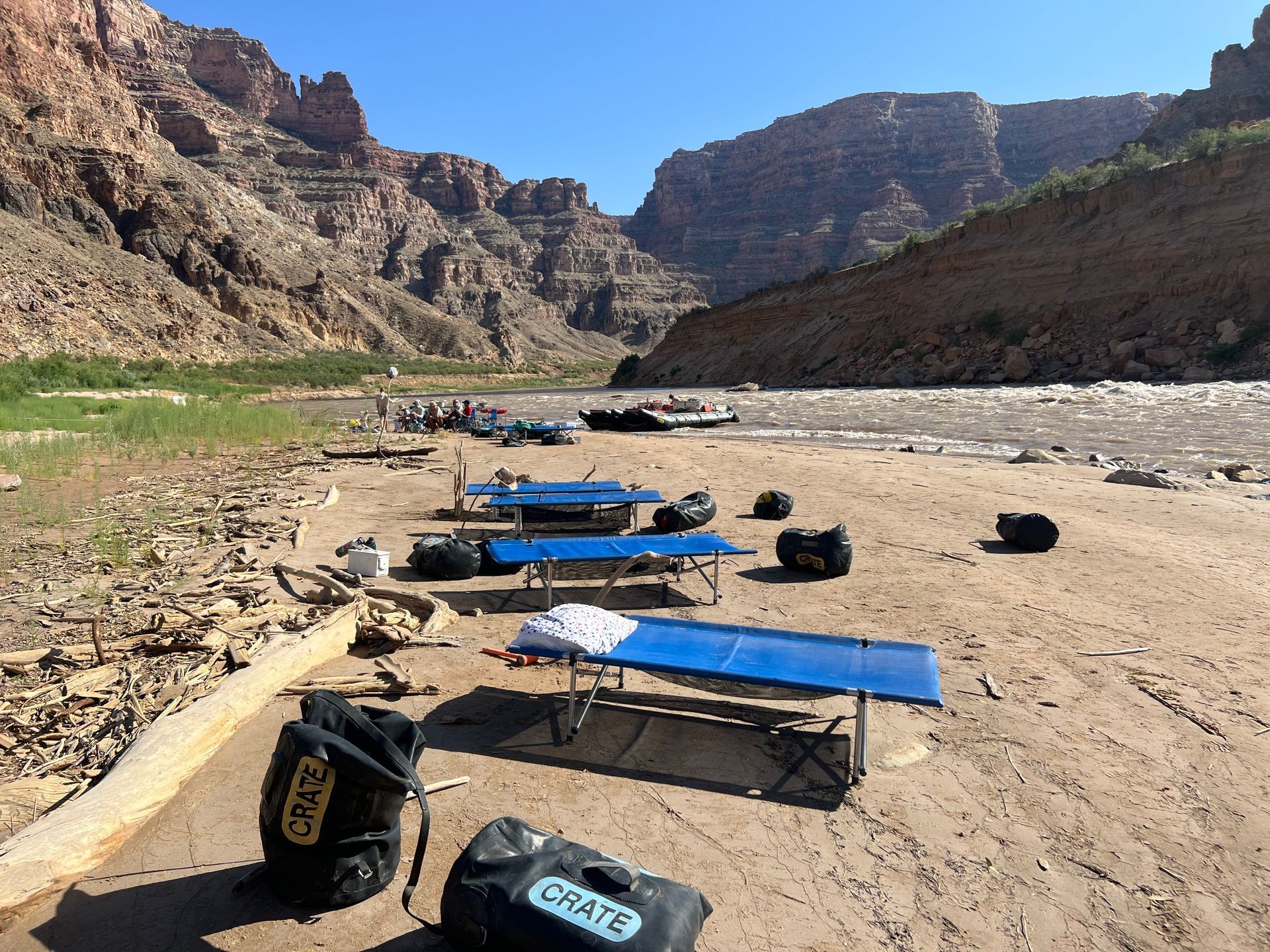 Rafting camp on a sandy riverbank in a canyon; blue cots, gear bags, and boats are present.