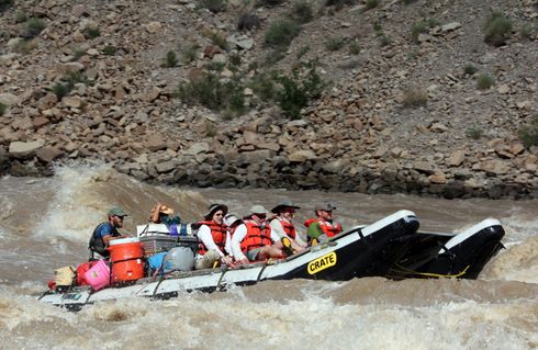 Raft with people navigating a whitewater rapid; rocky canyon walls in background.
