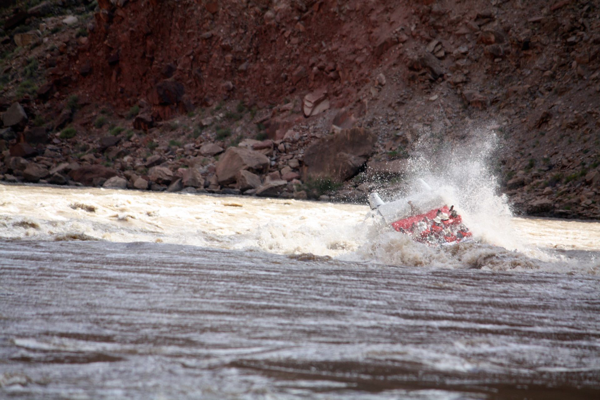 A raft capsizes in a turbulent river, spraying water against a red-brown rock face.