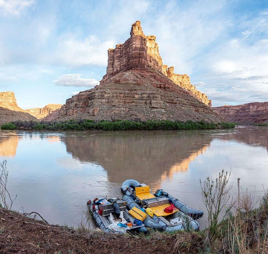 Inflatable rafts on river in front of towering brown rock formation under cloudy sky.