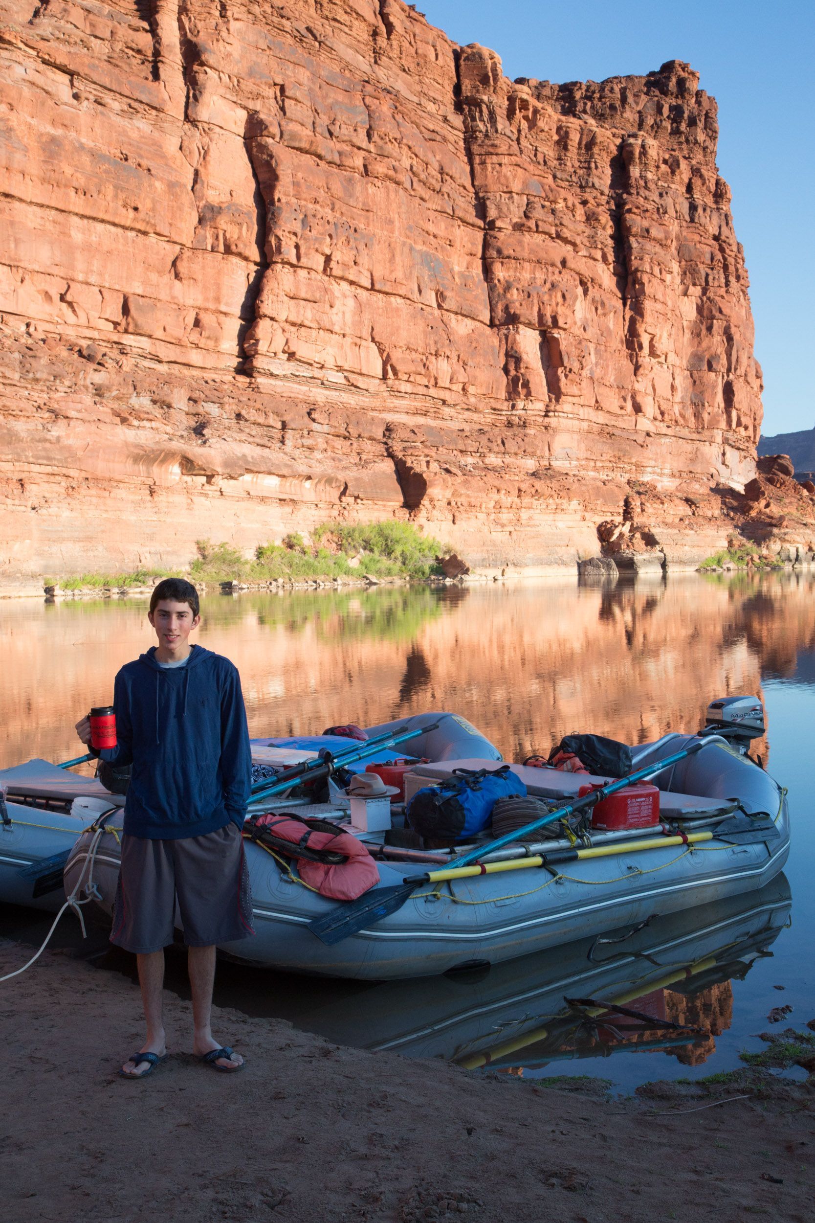 Man standing near rafts on a riverbank, red rock cliff backdrop.