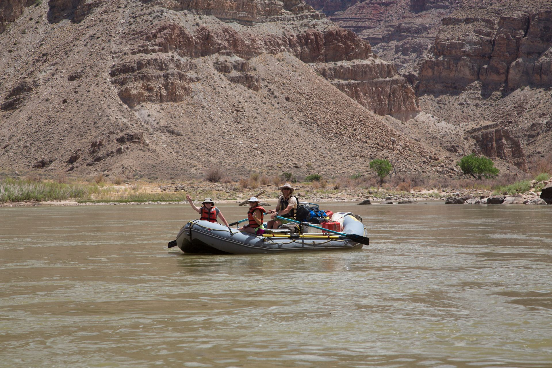 Rafting on a brown river, with passengers and gear, against a backdrop of tan canyon walls.