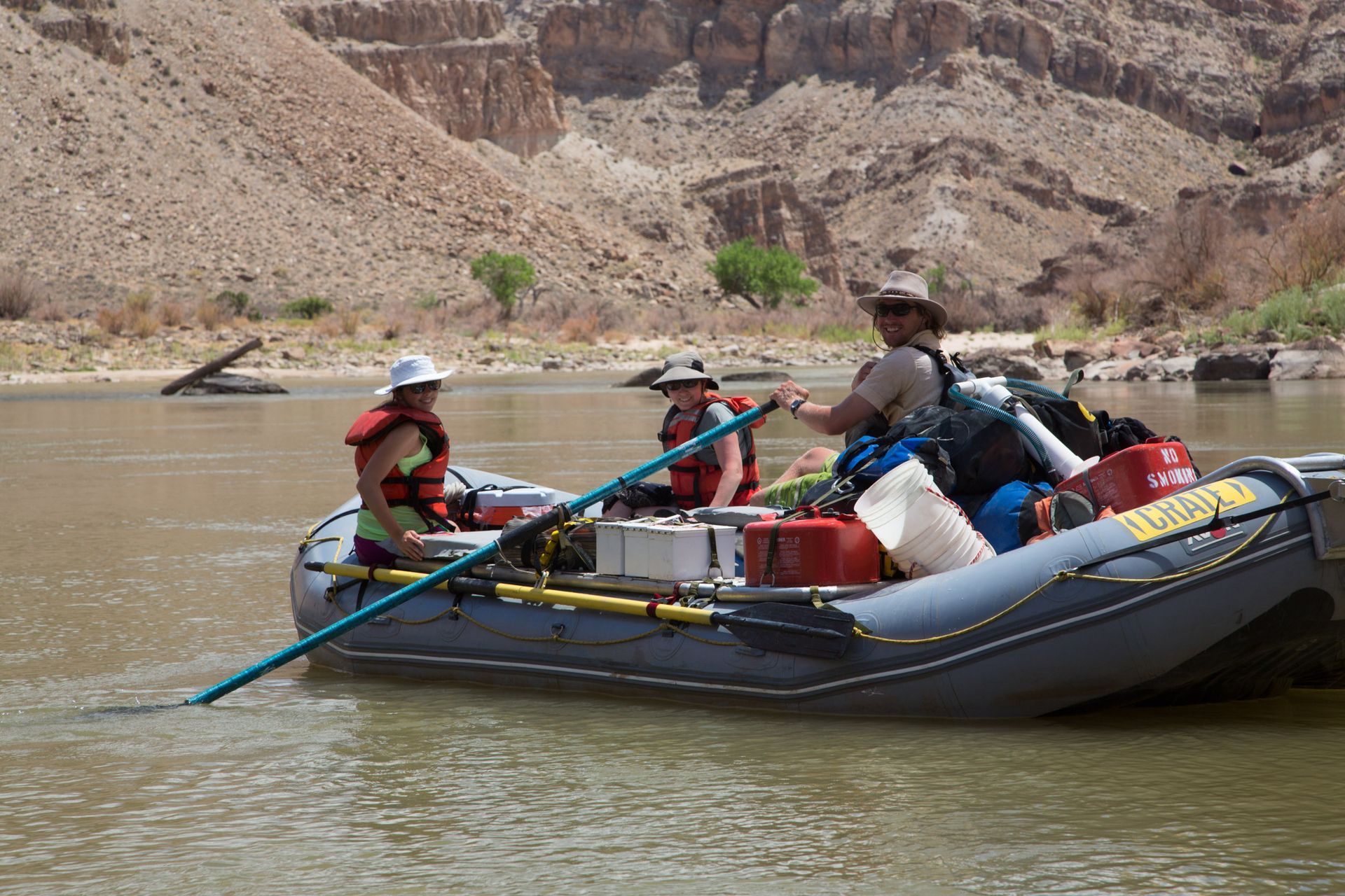 Three people on a raft in a river, using poles to navigate near a brown canyon.