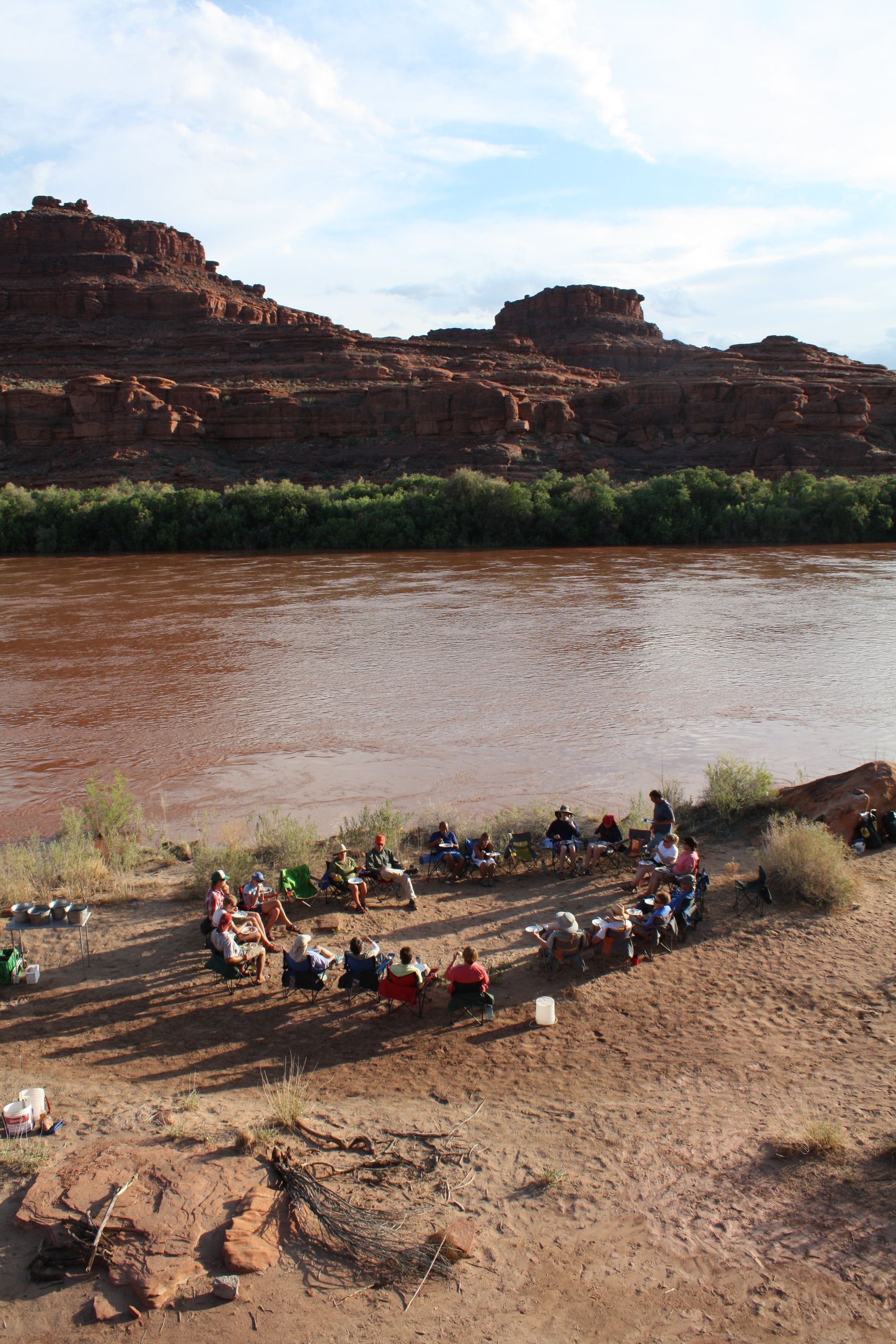 Group of people seated in a circle on riverbank; red cliffs and muddy river in background.