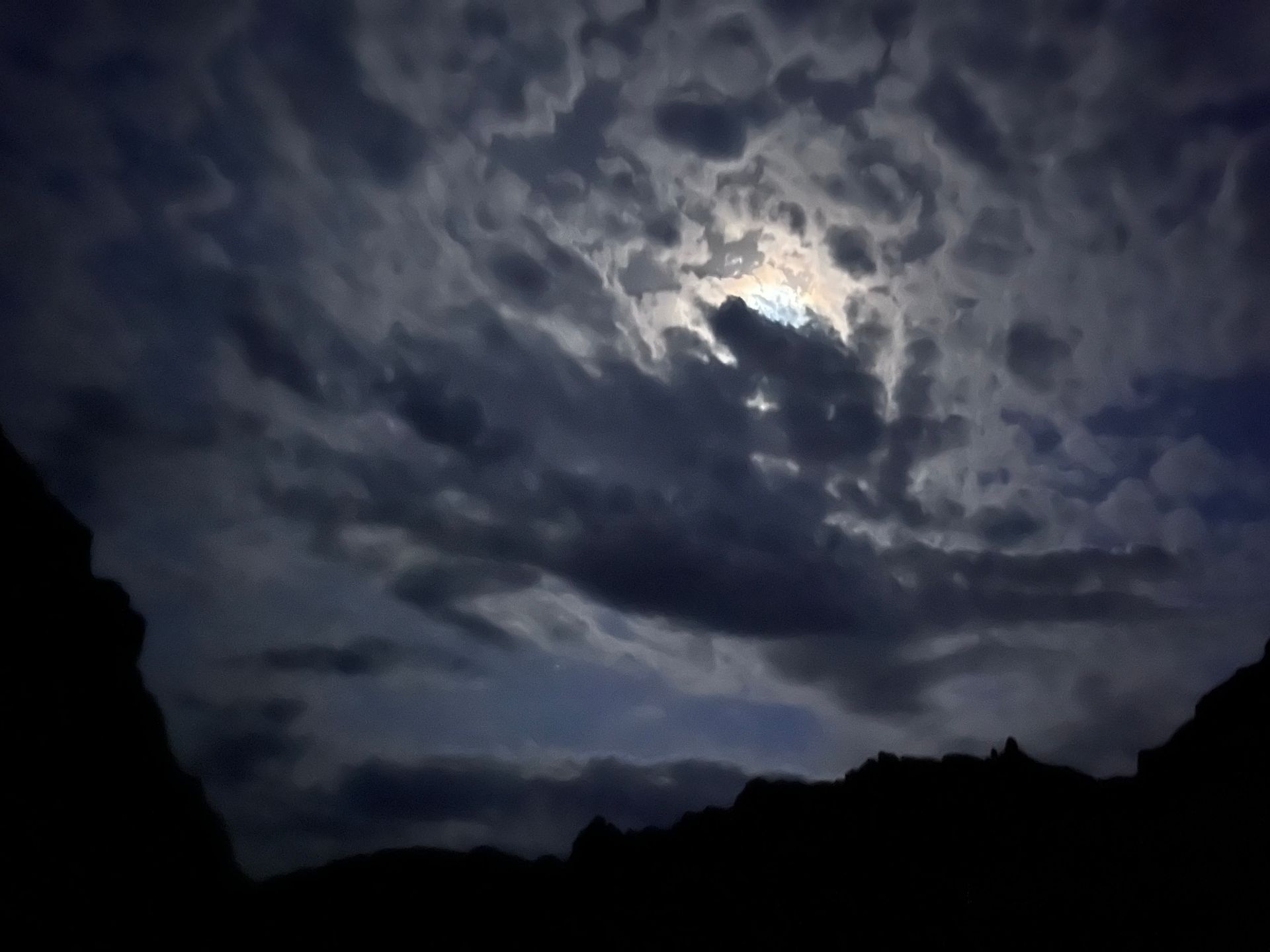 Dramatic night sky with swirling, dark clouds obscuring a bright moon, silhouetted mountains below.