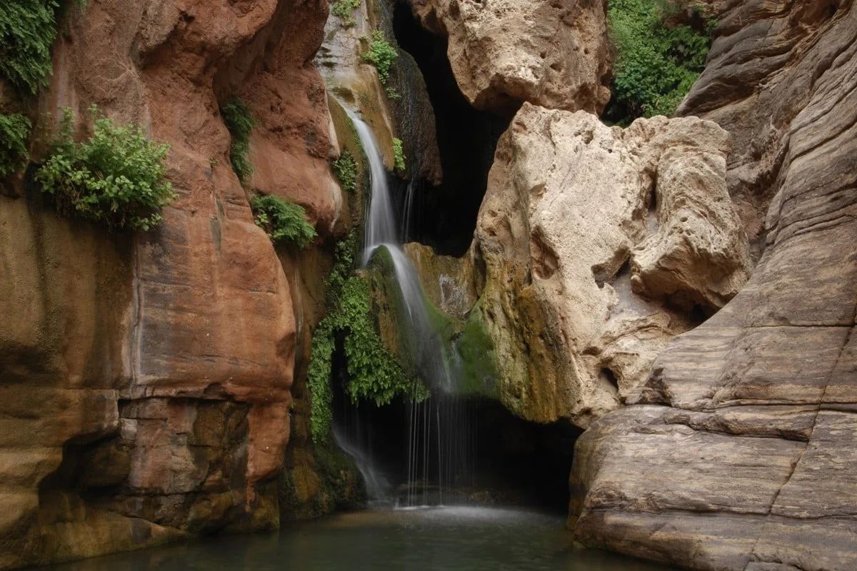 Waterfall cascading into a small pool, surrounded by layered, tan rock formations with green vegetation.