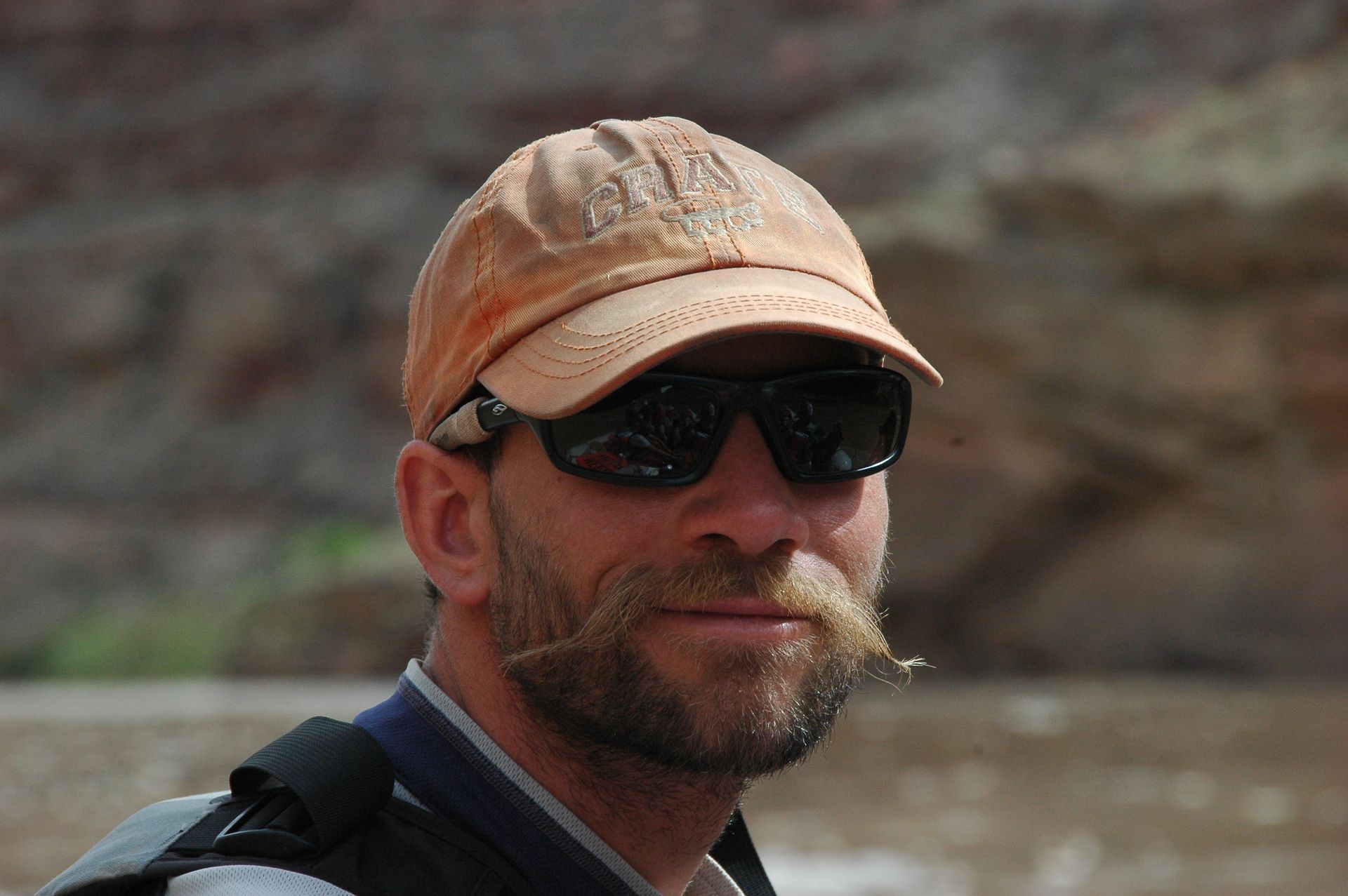 Man with mustache and sunglasses wearing cap, set in outdoors.