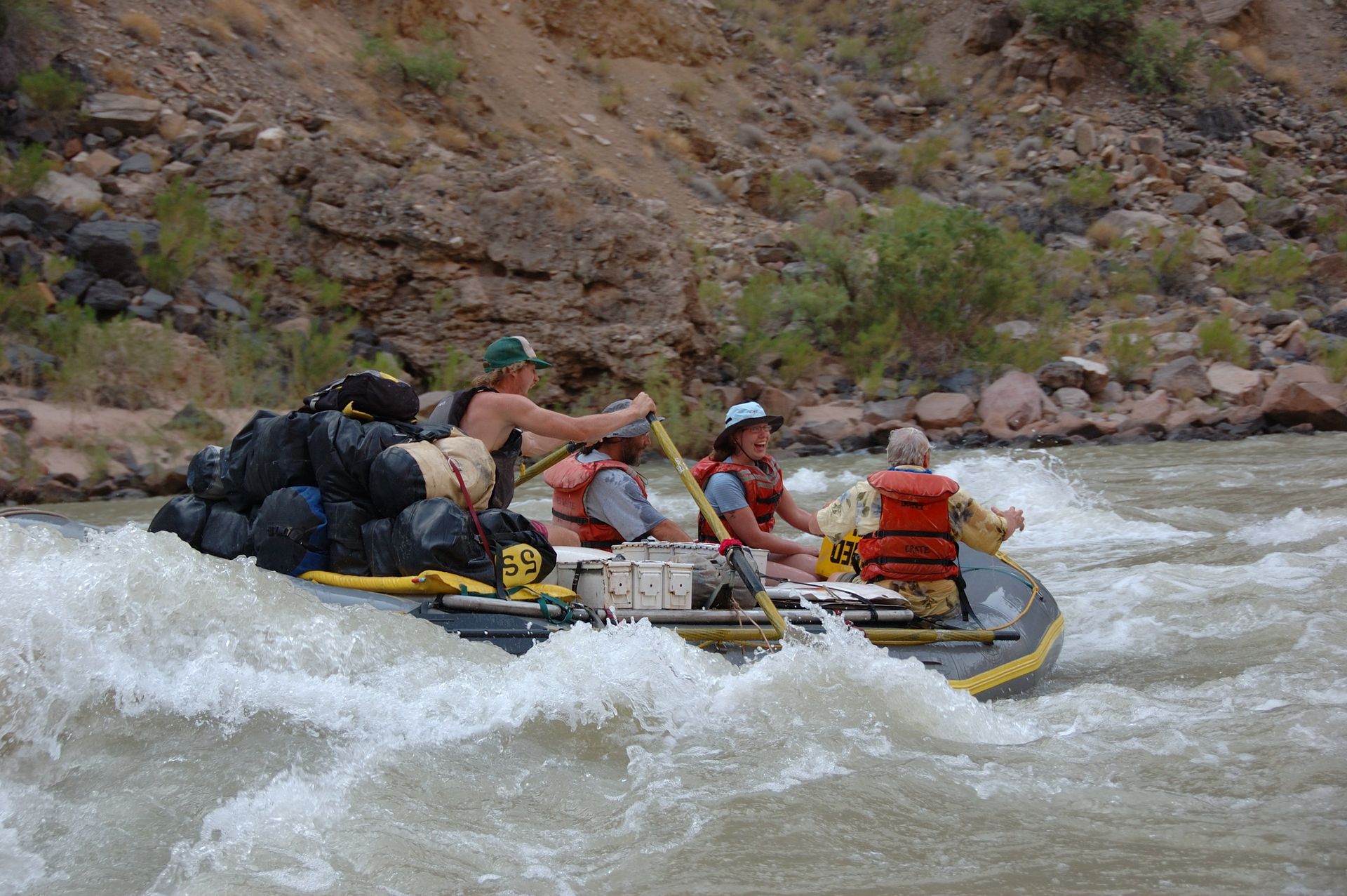Rafting through whitewater rapids; river surrounded by rock cliffs. People paddling, gear piled on raft.