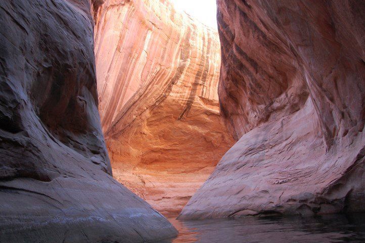 Narrow slot canyon with water, displaying orange and tan rock formations.