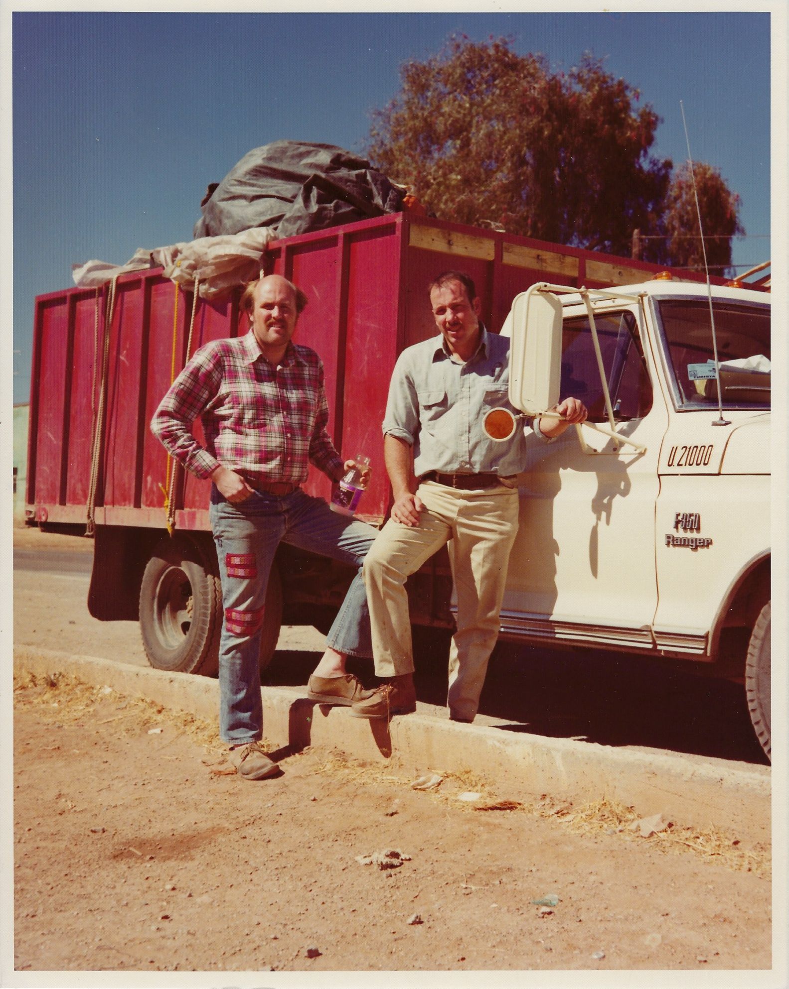 Two men pose next to a red truck loaded with goods. One leans on the cab; the other stands nearby.