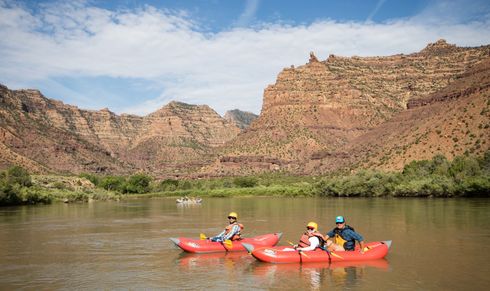 People in two red inflatable kayaks on a river, with red rock canyon in background.