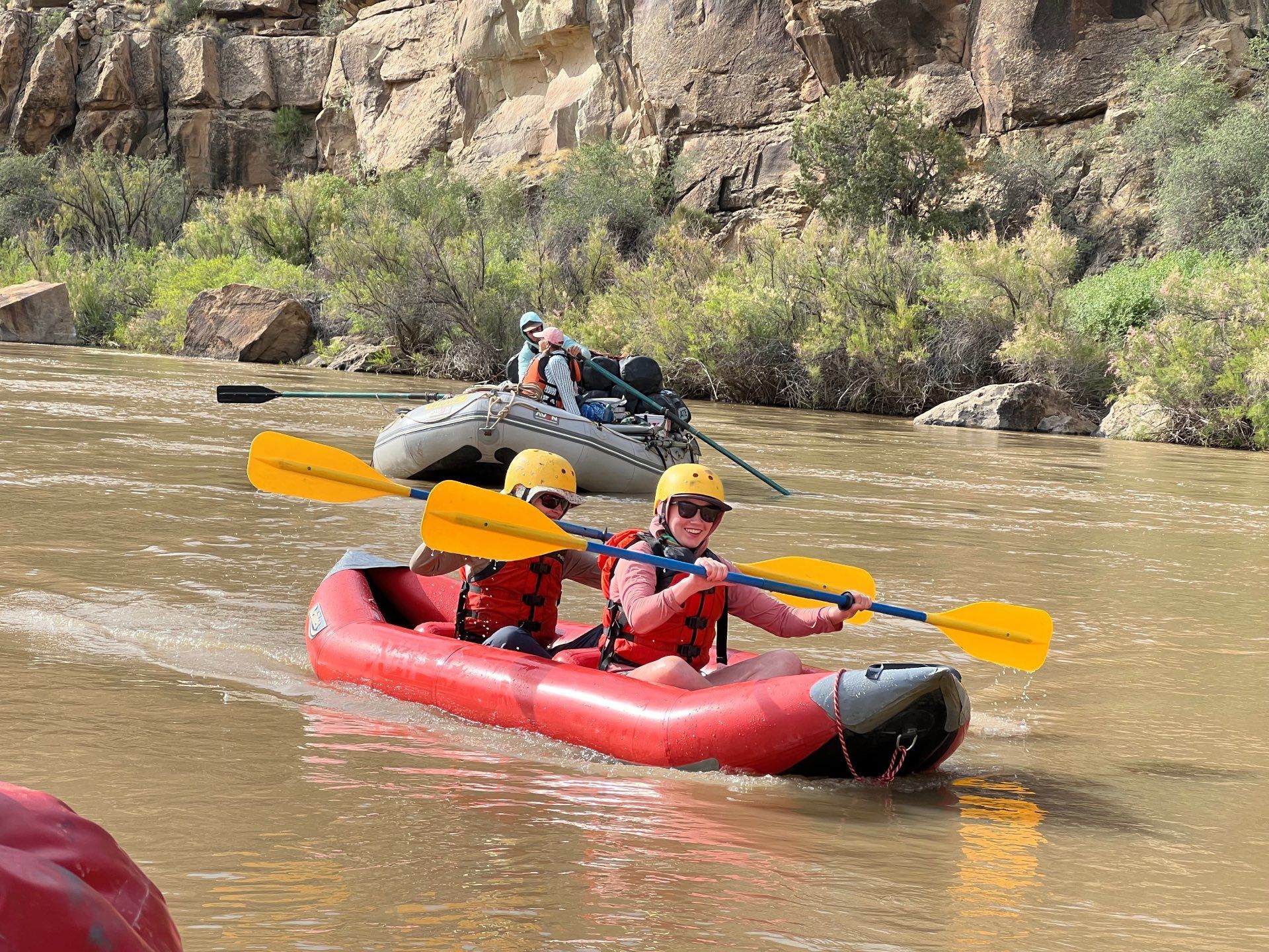 Two people on a raft in a canyon river, wearing life vests and hats.