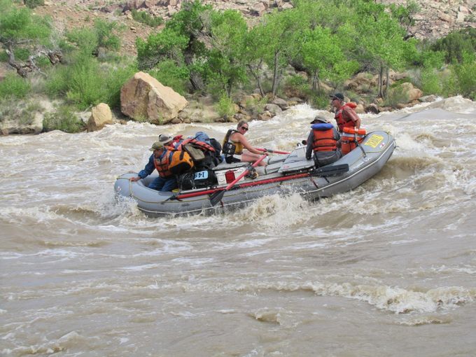Raft navigating a rapid on a brown river. Passengers wear life jackets, some paddling. Rocky banks in background.