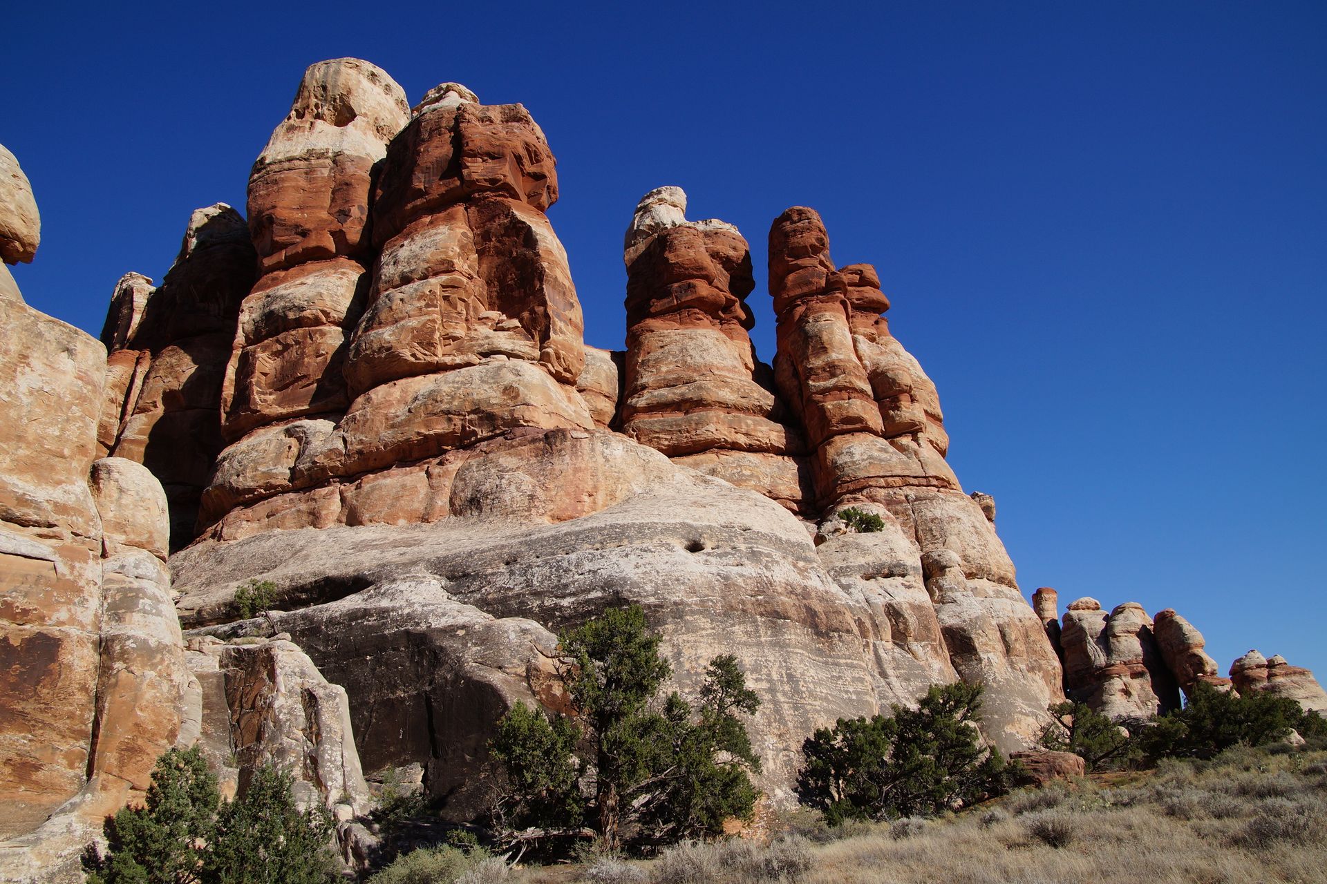 Red and tan rock formations against a bright blue sky, with sparse green shrubbery.