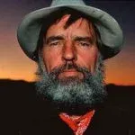 Man with a gray beard and hat, wearing a red scarf, with a sunset background.