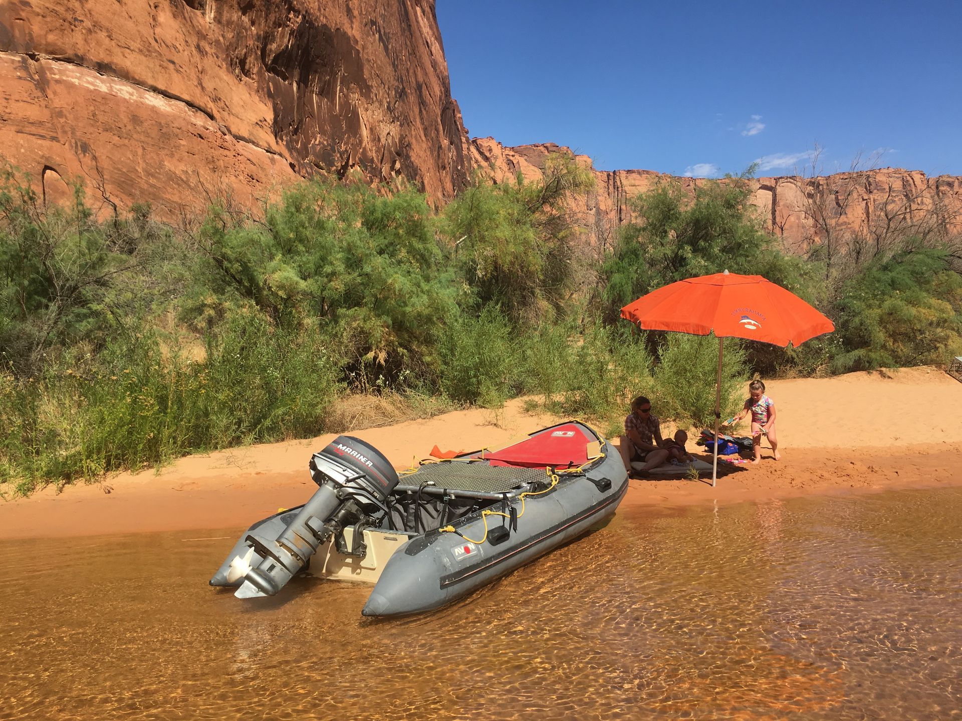 Sandy beach inside a cavern overlooks a river, with red rock canyon walls and a blue sky.