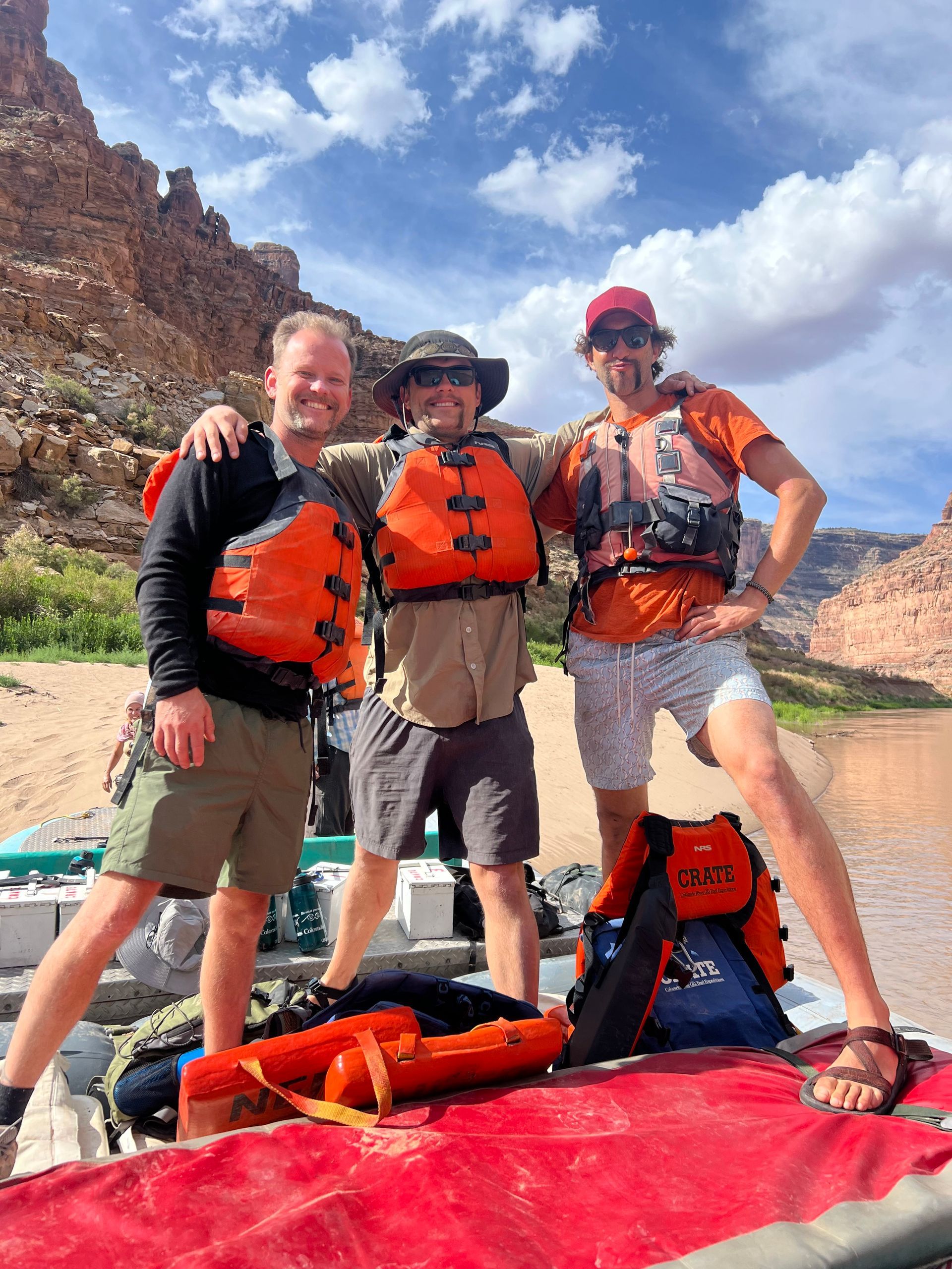 Three people in life vests on a raft in a river, posing with arms around each other, sunny day with canyon walls.
