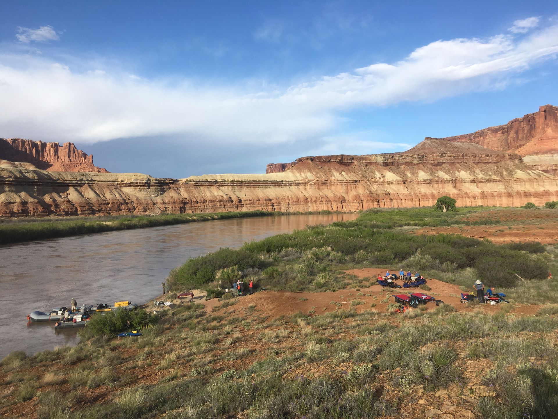 River with rafts, campers, and red rock cliffs under a partly cloudy blue sky.