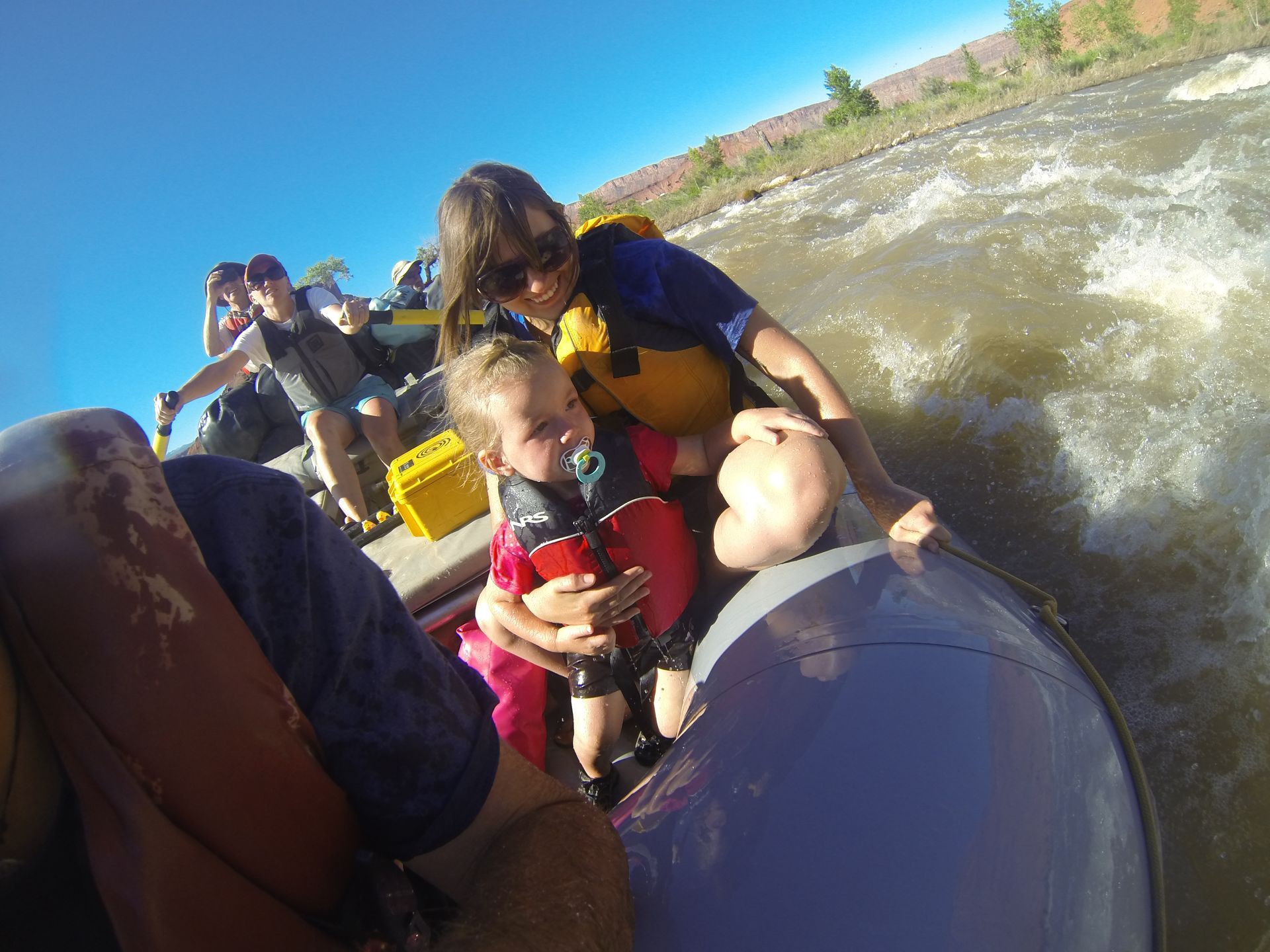 Woman holding a baby on a rafting boat; the baby is wearing a life jacket and has a pacifier.