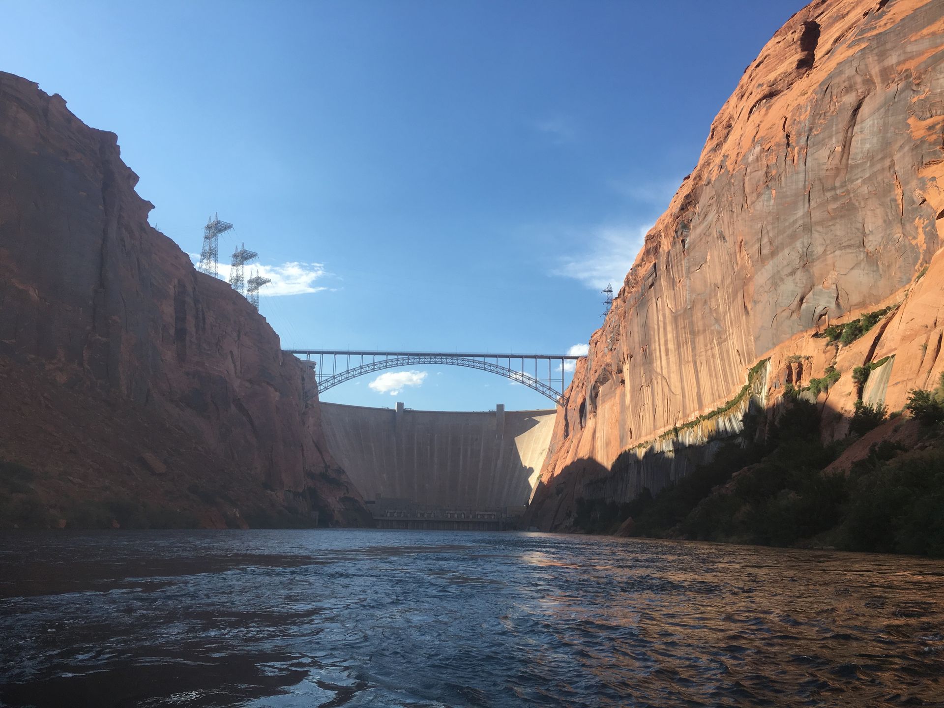 Glen Canyon Dam with bridge and canyon walls under a blue sky. Water flows towards the dam.