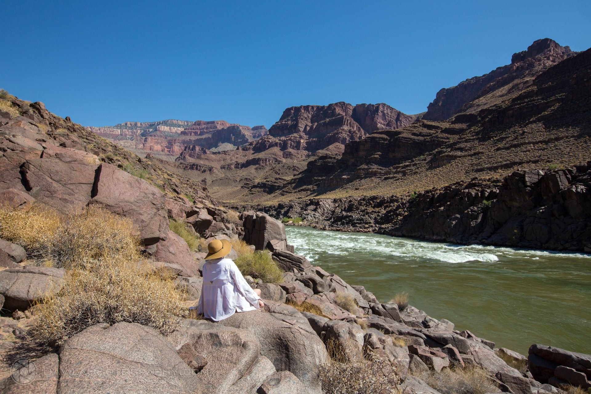 Person in white dress and hat sits by a rushing river in a canyon under a bright blue sky.