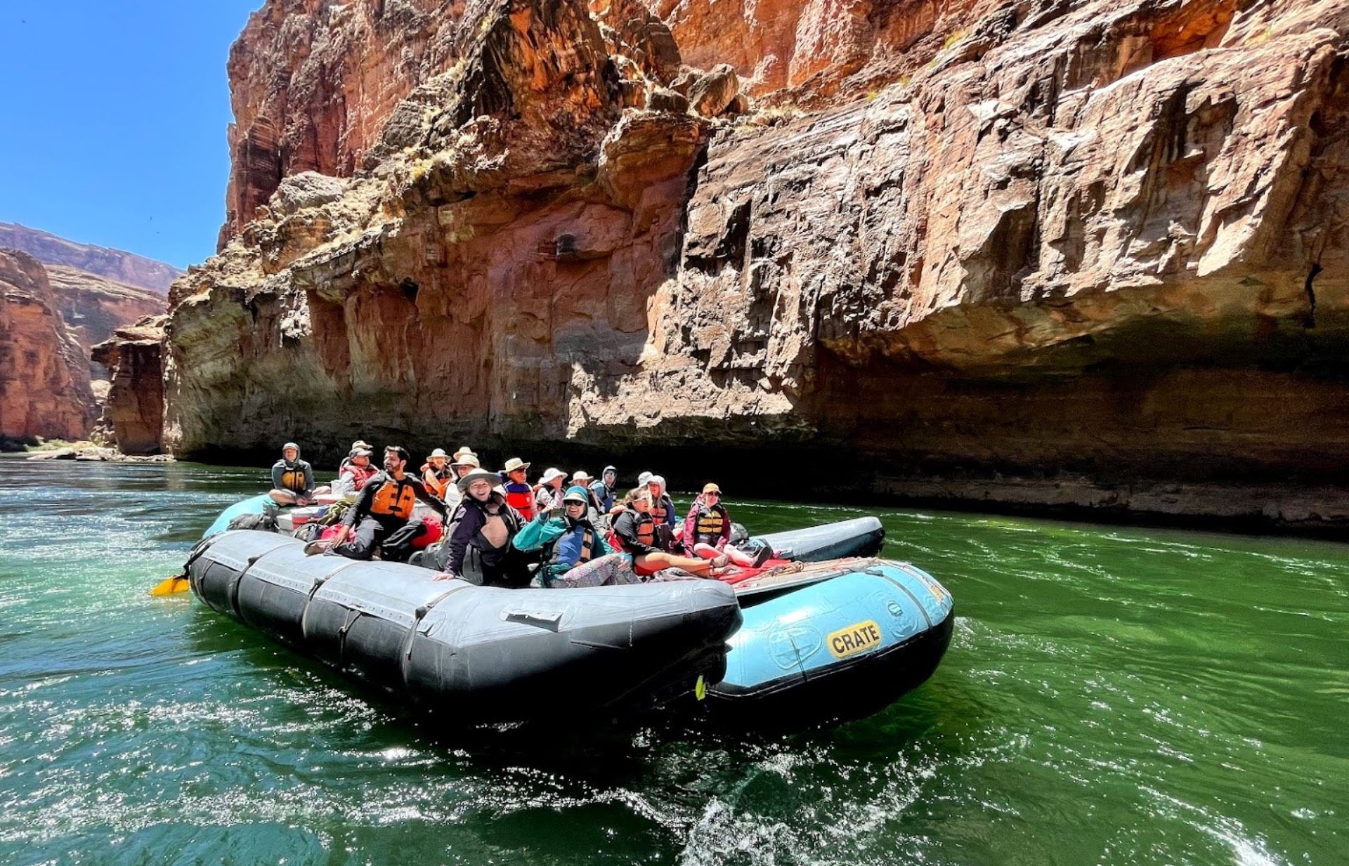 Raft with people on a green river, navigating through a canyon with brown rock walls under a blue sky.
