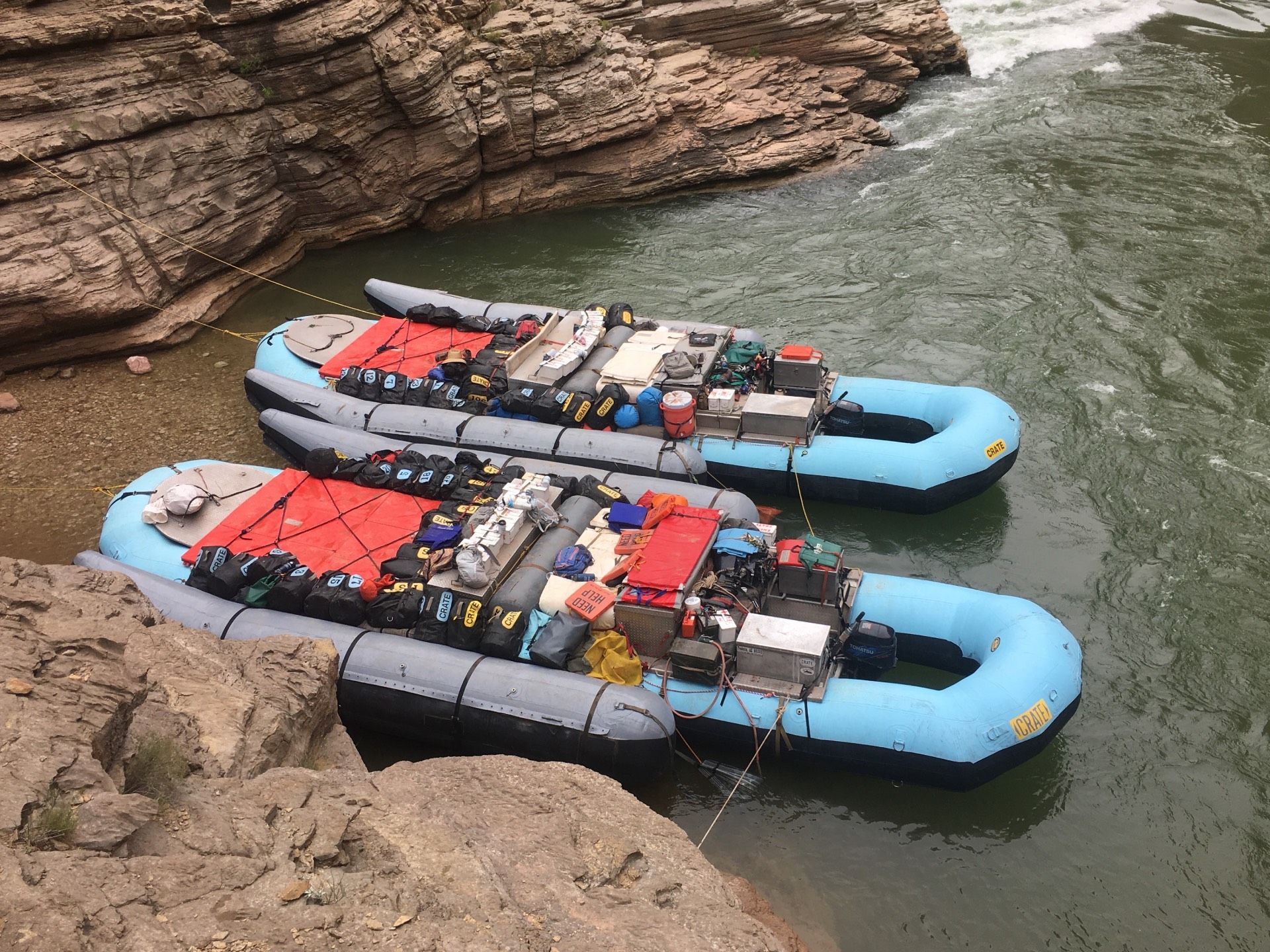 Two blue rafts loaded with gear on a river near a rocky bank.