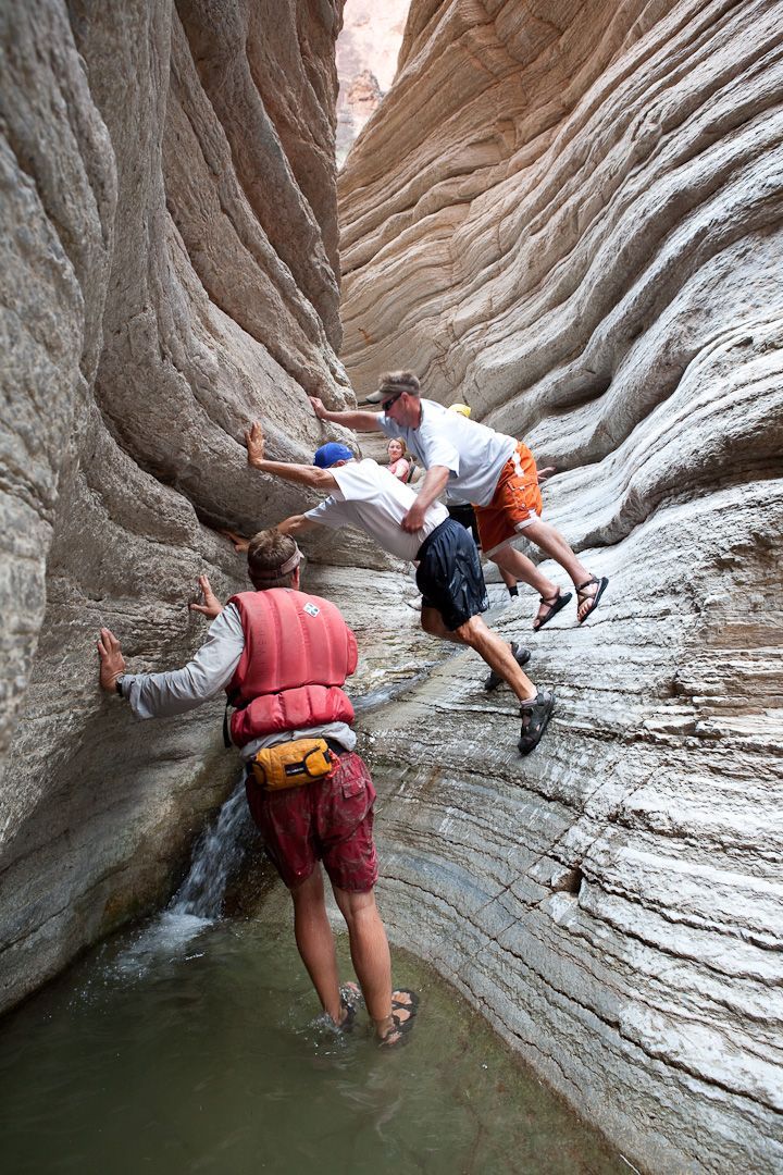 Three people climb out of a narrow canyon with layered rock walls. One wears a life vest, helping the others.