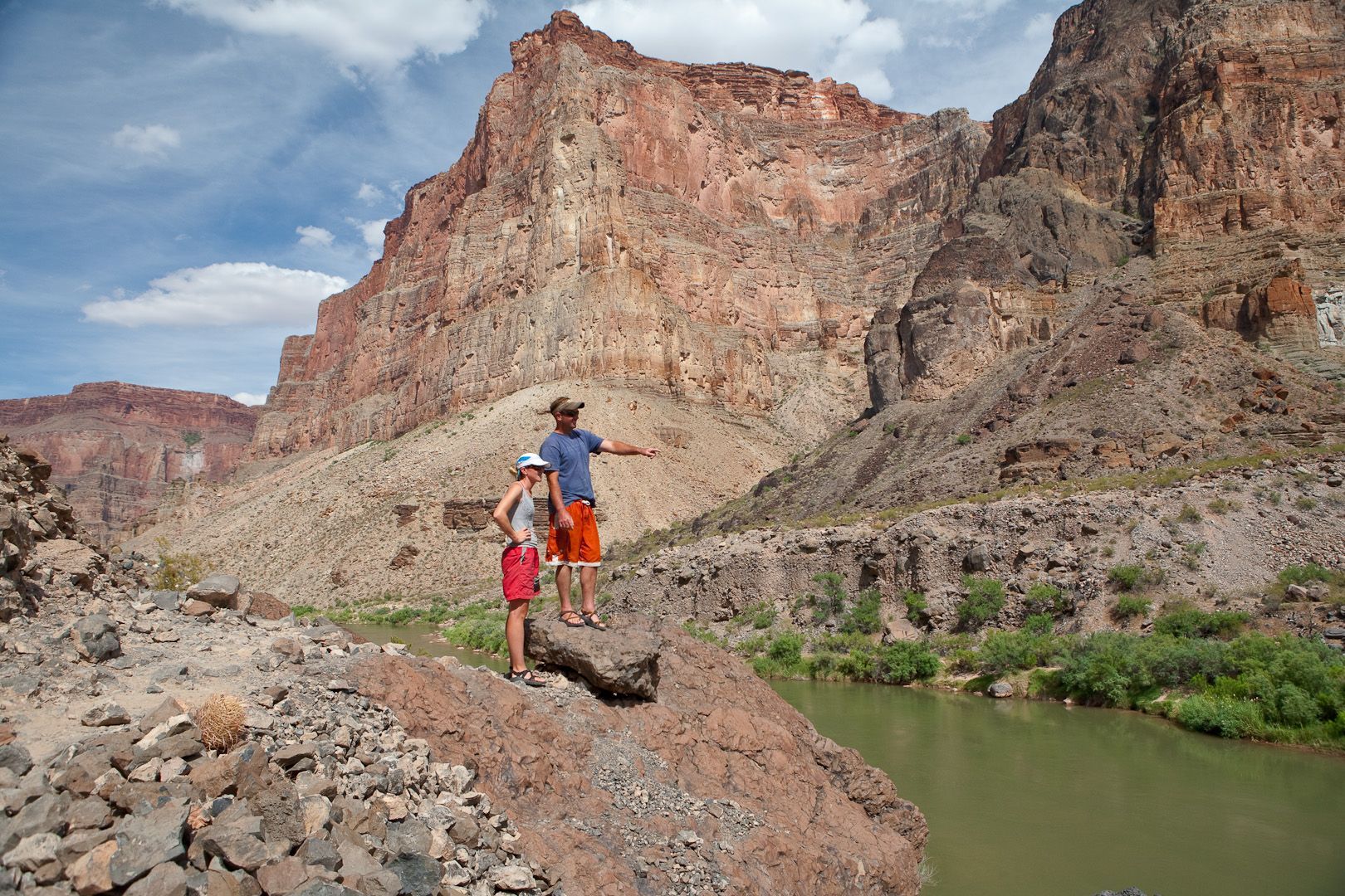 Two people standing by a river, pointing at a red canyon wall.