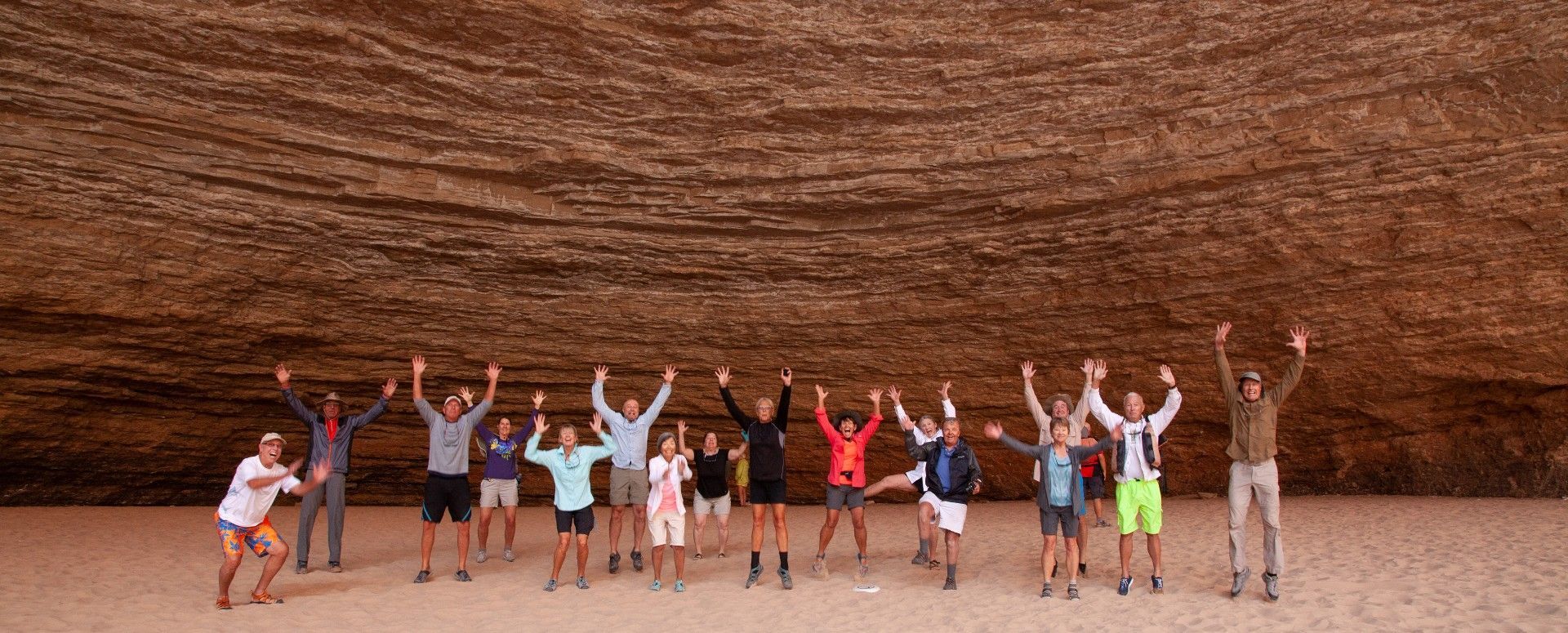 Group of people with arms raised in front of a layered rock formation. Sandy ground.
